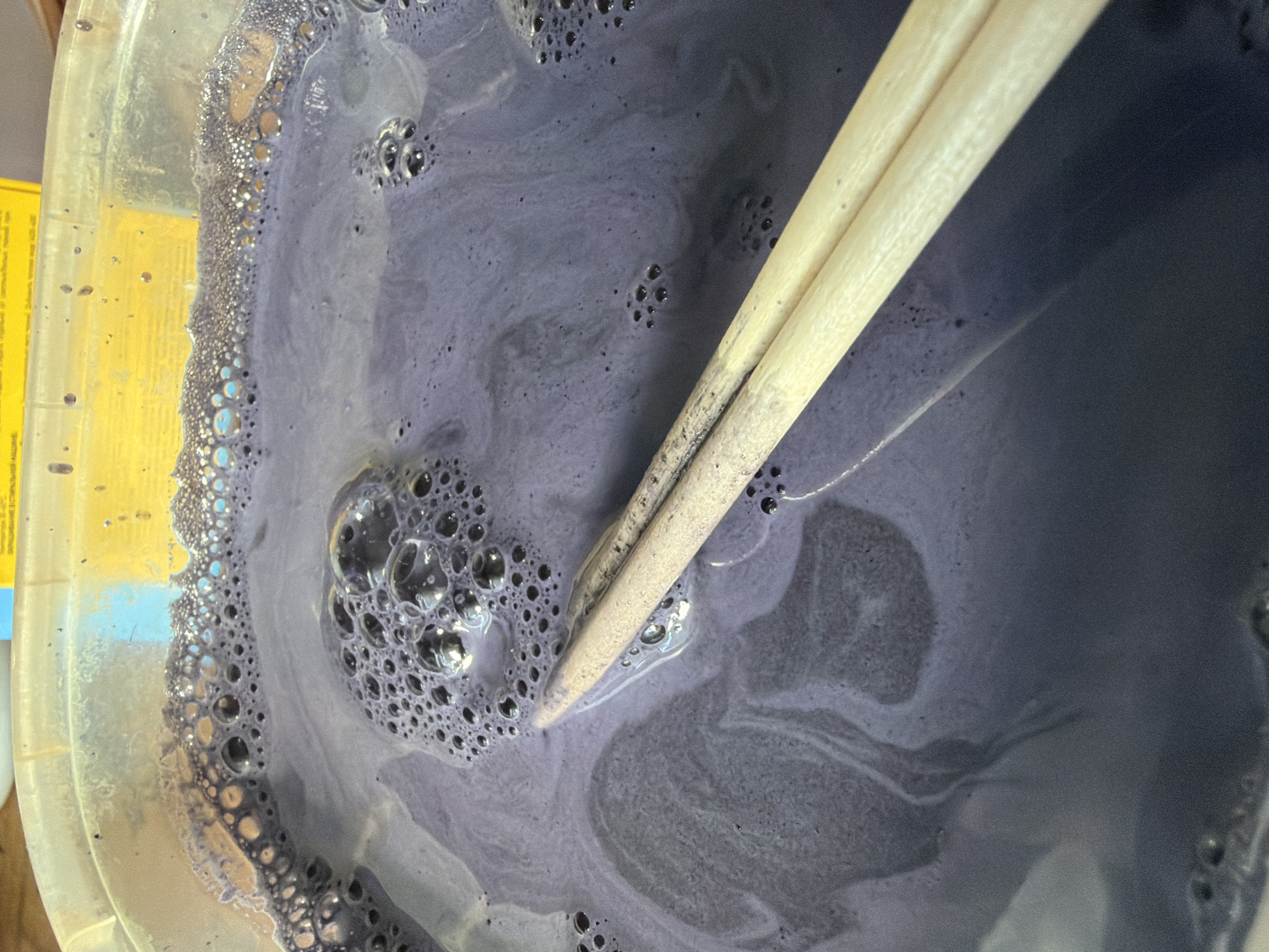 The image shows a close-up view of a plastic container filled with a dark purple liquid that has a foamy surface. Two light-colored wooden chopsticks are submerged in the liquid, positioned diagonally across the frame. The liquid appears to be some sort of dye or coloring solution, possibly for fabric or art. The container's rim is visible at the top, and a yellow label with Russian text is partially visible behind it. The scene suggests a creative or crafting activity. There is no discernible indication of a specific location within the city of Chaikovsky, Russia.