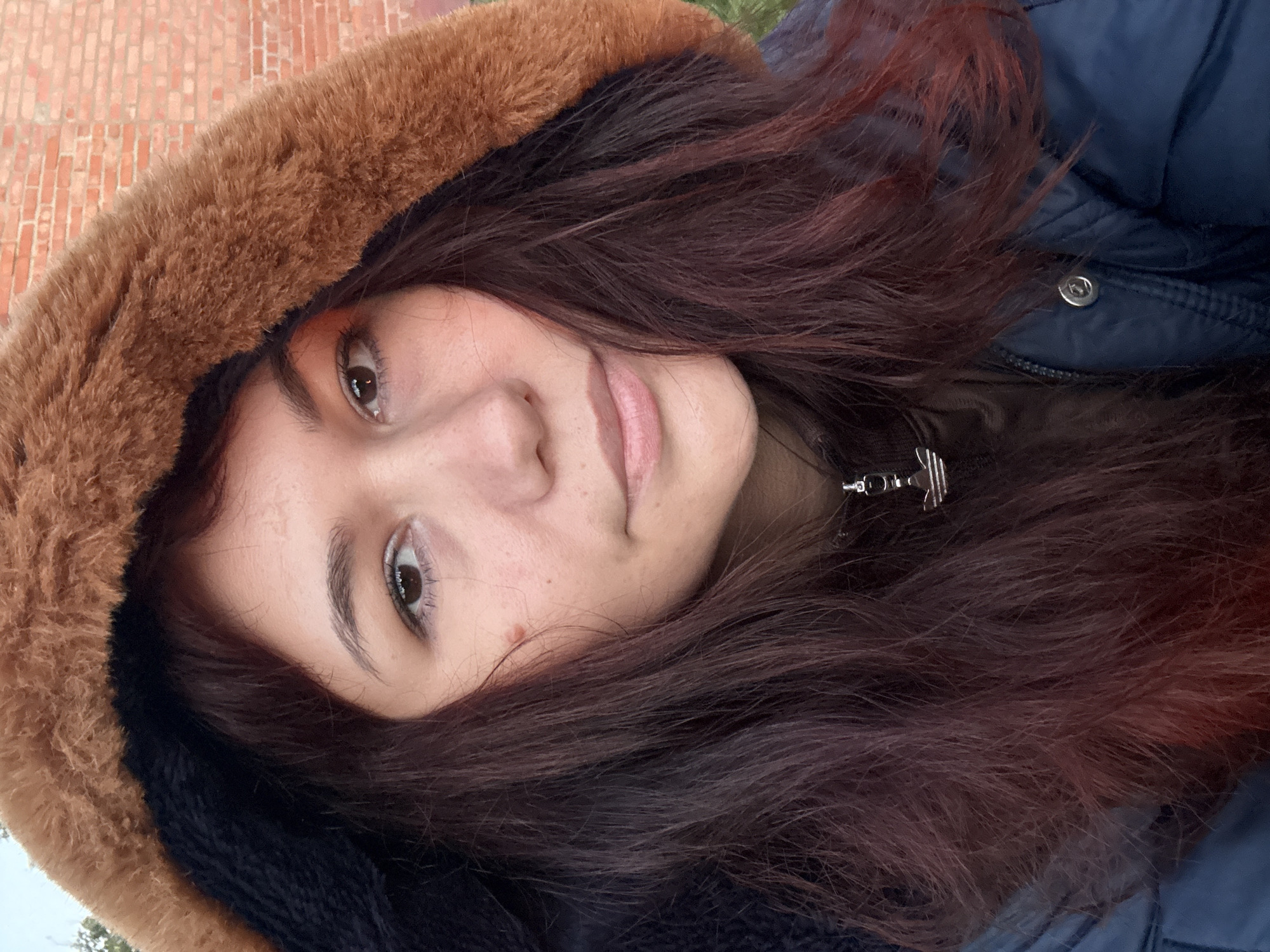The image is a close-up selfie of a young woman with dark red hair. She is wearing a dark blue puffer jacket with a brown faux fur-lined hood. Her hair is loose and falls around her face and shoulders. She is wearing light makeup, including eyeshadow with an orange hue. A silver pendant with the Adidas logo hangs from a chain around her neck. The background is slightly blurred but appears to be an outdoor setting with a brick wall visible behind her on the right. The lighting suggests it is daytime. The overall impression is a casual, personal photograph. There is no indication that the image was taken in Imelevo, Russia.