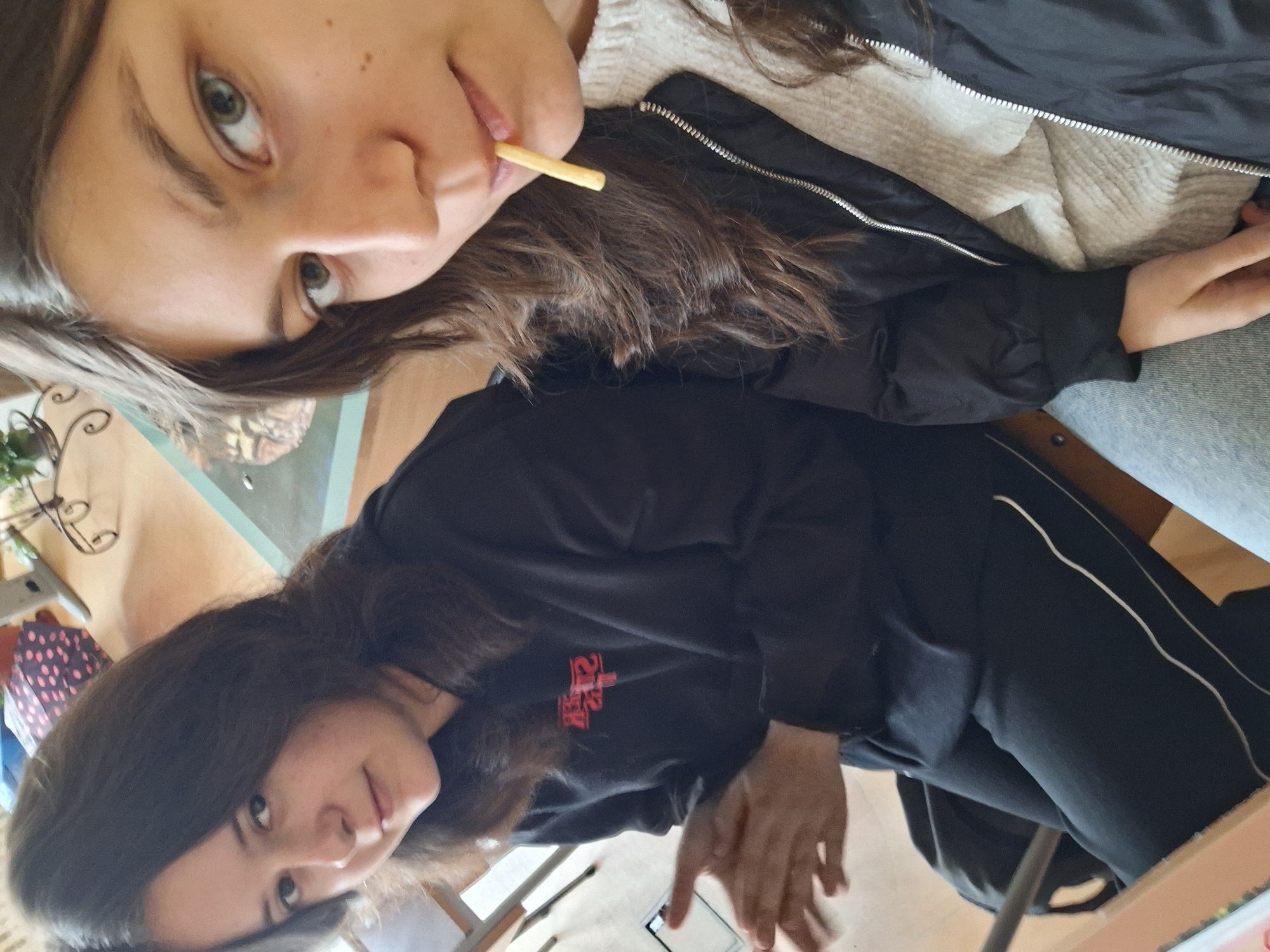 Two young women are seated at a table indoors. The first woman, in the upper part of the image, is wearing a black jacket and has a french fry in her mouth. The second woman, in the lower part of the image, is wearing a black sweater with a red logo and black pants. The setting appears to be a cafe or restaurant, indicated by the table, chairs, and items like a potted plant and polka-dot patterned bag visible in the background. The location is Panagyurishte, Bulgaria.