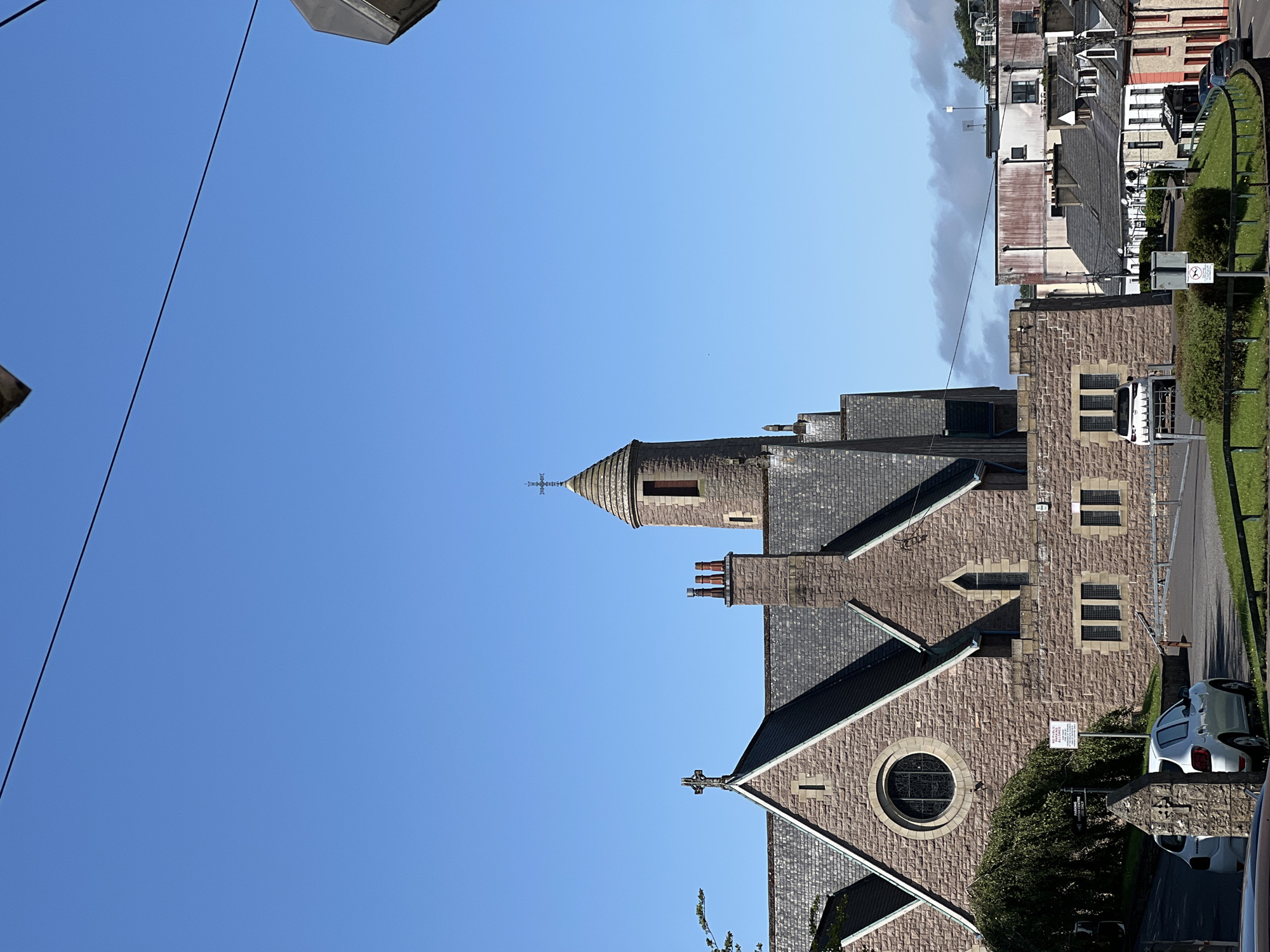 The image captures a view of a stone church with a prominent tower topped by a spire. The church features a circular stained-glass window and several smaller rectangular windows. Adjacent to the church are other buildings and houses, indicating a built-up area. A white car is parked in front of the church, and a metal barrier is visible on a road. The sky is clear and blue, with some wispy clouds in the distance. The scene suggests a daytime setting in a town or city. The presence of the church and the architecture of the surrounding buildings hint at a location with historical significance, possibly in Ireland.