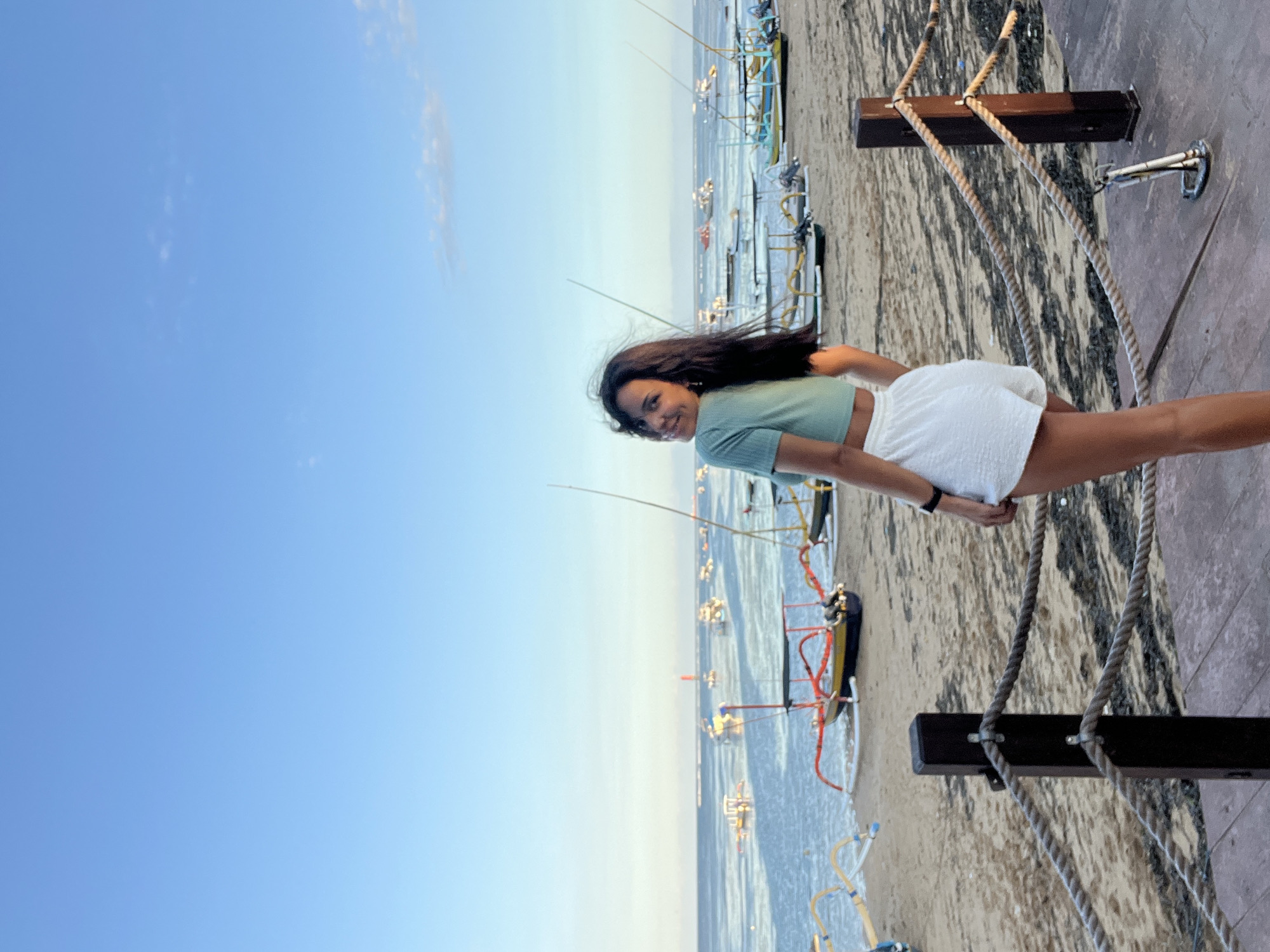 A young woman is standing on a wooden boardwalk next to a beach, with the ocean and sky behind her. She is wearing a light green short-sleeved crop top and white shorts. She is looking and smiling slightly at the camera. The ocean is calm, with many small boats docked in the distance. The sky is a light blue with a few wispy clouds. There are rope barriers and wooden posts on the boardwalk, and the beach is sandy. The location is in Denpasar, Indonesia, as evidenced by the outrigger boats visible on the beach.