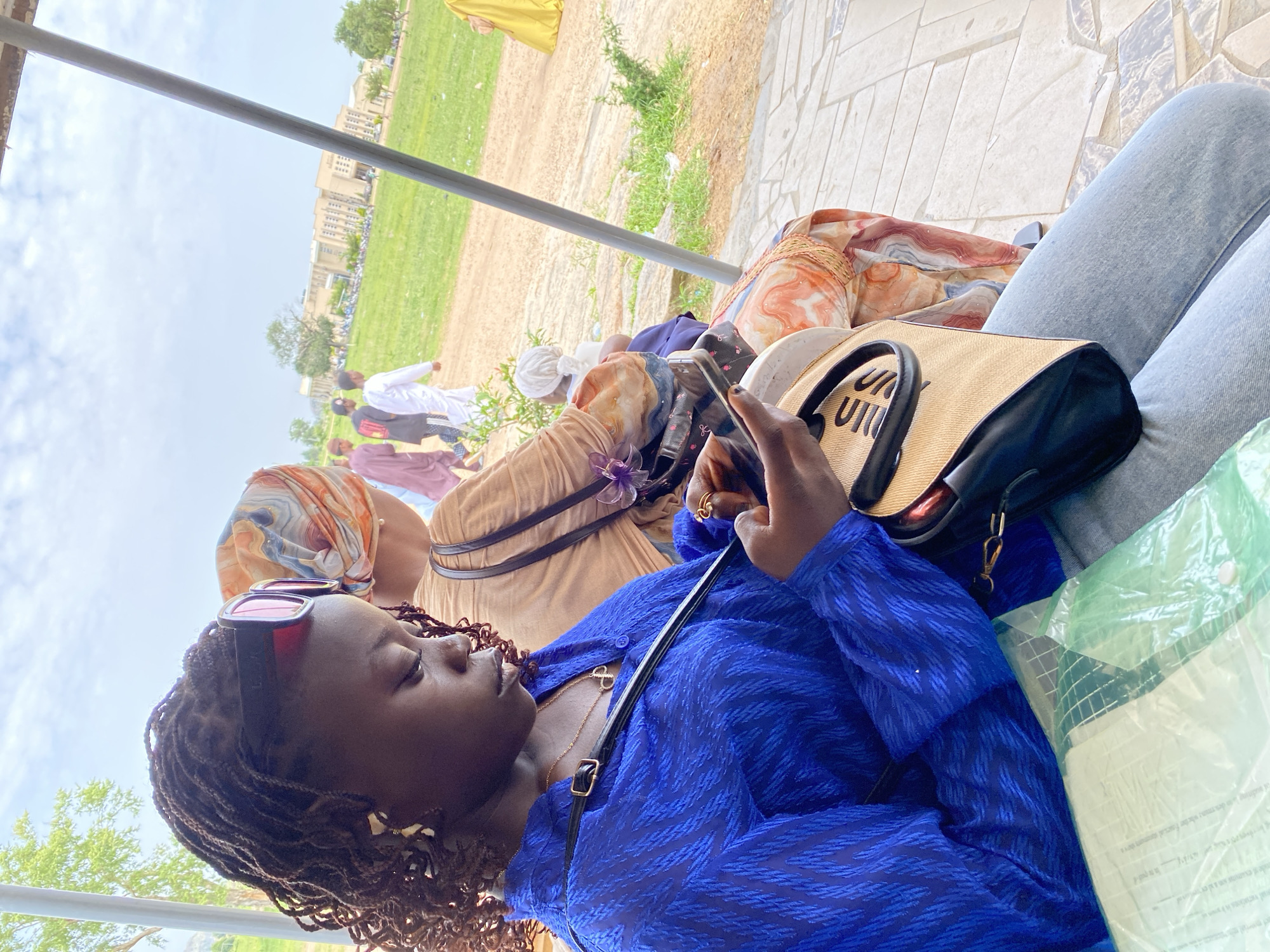 The image, taken in Yali, Nigeria, features a young woman in the foreground, seated and engrossed in her mobile phone. She wears a vibrant blue patterned shirt, a gold necklace, and pink-rimmed sunglasses pushed atop her braided hair. A beige and black handbag rests on her lap. Beside her, another woman is partially visible, wearing a beige top and an orange, blue, and white patterned headscarf and skirt. In the background, several individuals, some in traditional attire, walk across a grassy area towards a large, modern building. The setting appears to be outdoors, possibly near an educational institution, with a cloudy sky overhead.