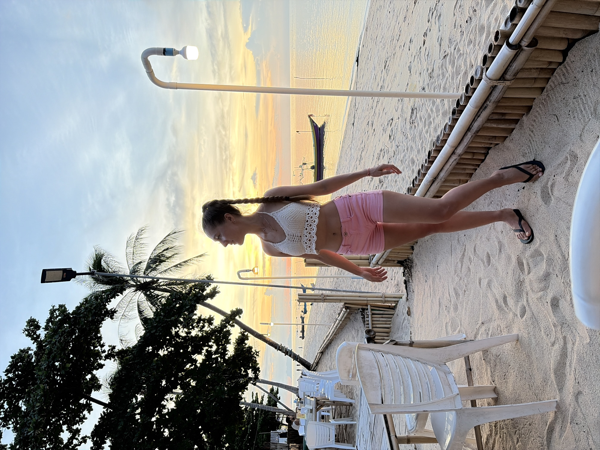 A woman with blonde hair pulled into a braid, wearing a white crocheted crop top and pink shorts, stands on a sandy beach. She is looking to her right, towards the ocean and the sunset. The beach has white plastic chairs and tables set up, suggesting an outdoor dining area. Palm trees and lush green foliage are visible to the left, and a wooden bamboo fence runs along the edge of the sand. Several boats are anchored in the calm water, and the sky is filled with soft orange and yellow hues of the setting sun. The scene is set in Baan Thong Po, Thailand, a coastal location known for its beaches and relaxed atmosphere.