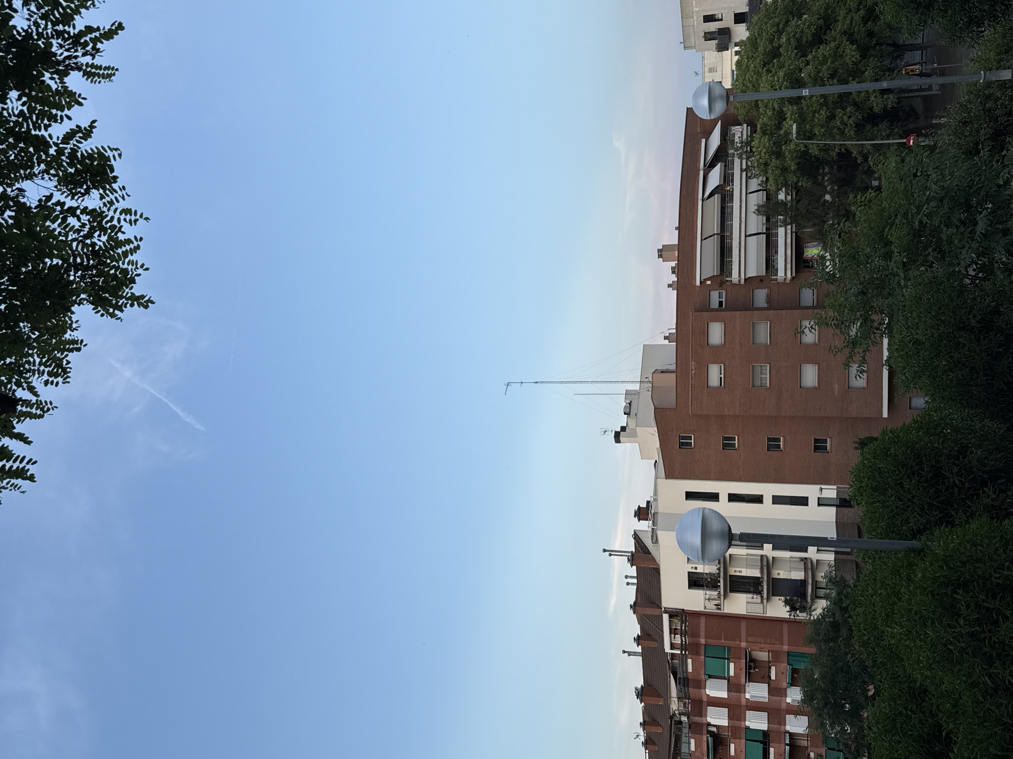 The image shows an exterior view of buildings and trees under a clear sky, likely in Barcelona, Spain. The sky is a pale blue, suggesting daytime, possibly late afternoon or early morning, with a faint contrail visible. Lush green trees flank the left and right sides of the frame, obscuring parts of the buildings and the sky.
The buildings are multi-story structures, with visible brick facades and numerous windows. Some buildings have balconies with white awnings. A street lamp with a spherical, metallic shade stands in front of one of the buildings. Another building, taller and lighter in color, is visible behind it. The rooftops show various elements like chimneys and antennas, indicating an urban setting. There is no visible text in the image. The overall scene depicts a residential or mixed-use urban environment.