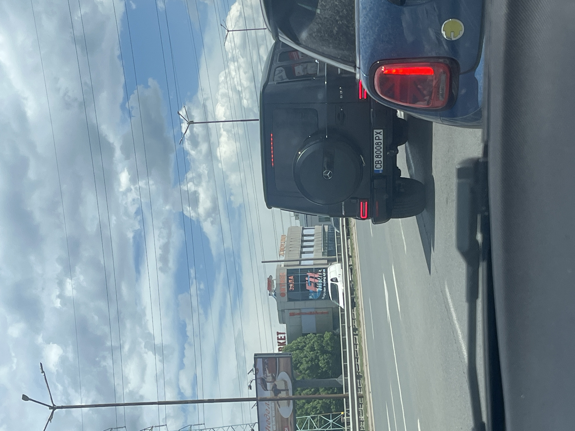 The image depicts a traffic scene in Sofia, Bulgaria. A black Mercedes-Benz G-Wagon is in front, identified by the license plate "CB 8008 PX." A blue car is partially visible to the right. The scene is viewed from within another vehicle, through its window. The roadside includes power lines and buildings, one with signs and posters written in Bulgarian. Visible commercial signs may include "Fit нес" and "MARKET." The sky above is partially cloudy.
