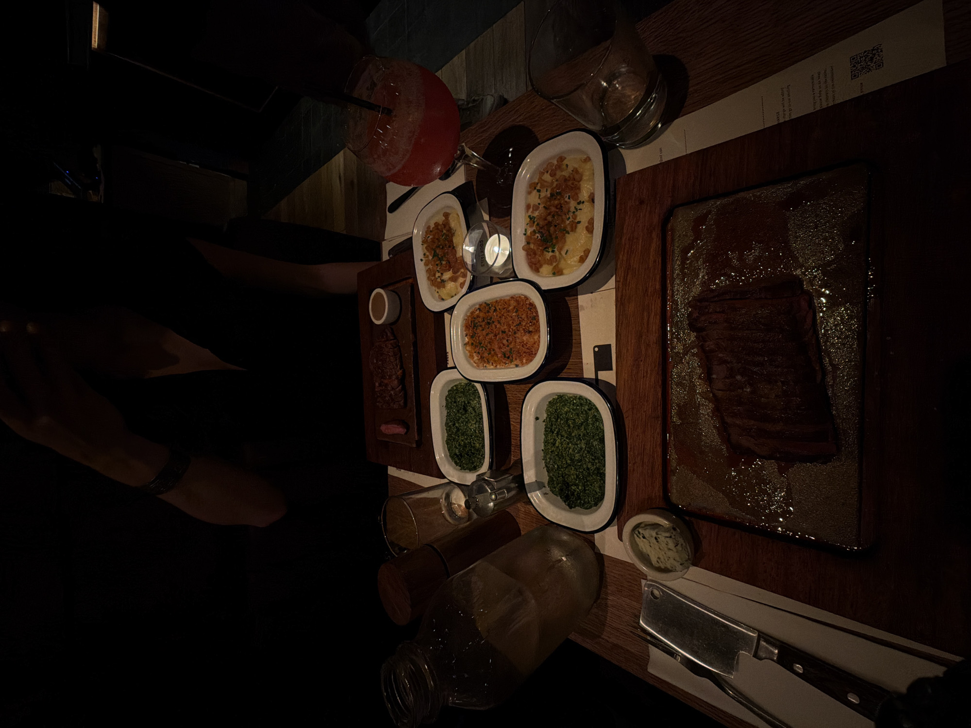 The image depicts a dimly lit dining table at what appears to be a restaurant, capturing an intimate evening meal. The scene is dominated by a dark wooden table laden with food and drinks, illuminated primarily by a small, lit candle in a glass holder.
The main dish is a large piece of grilled meat, likely steak, presented on a rectangular serving board that has a glossy, wet appearance, possibly from cooking juices. Surrounding it are several white, oval-shaped dishes containing various side dishes: two appear to be creamy potatoes or mac and cheese topped with crispy fried onions, one holds a reddish-orange granular dish, and two contain vibrant green creamed spinach.
Beverages include a red cocktail with a straw in a large glass, an empty glass pitcher, and a clear glass bottle. A small white ramekin with butter or sauce is also present. Tableware includes a prominent, heavy cleaver-style knife with a dark wooden handle, resting on a white napkin next to a fork.
Partial views of at least two people are visible in the low light, with an arm wearing a dark bracelet noticeable on the left side of the frame. A paper menu or placemat is visible under some dishes, displaying a QR code and the word "cocktails," along with other indistinct text, including phrases like "cognac aged in" and "crispy bacon". The overall ambiance suggests a cozy, upscale casual dining experience, fitting for its London setting.