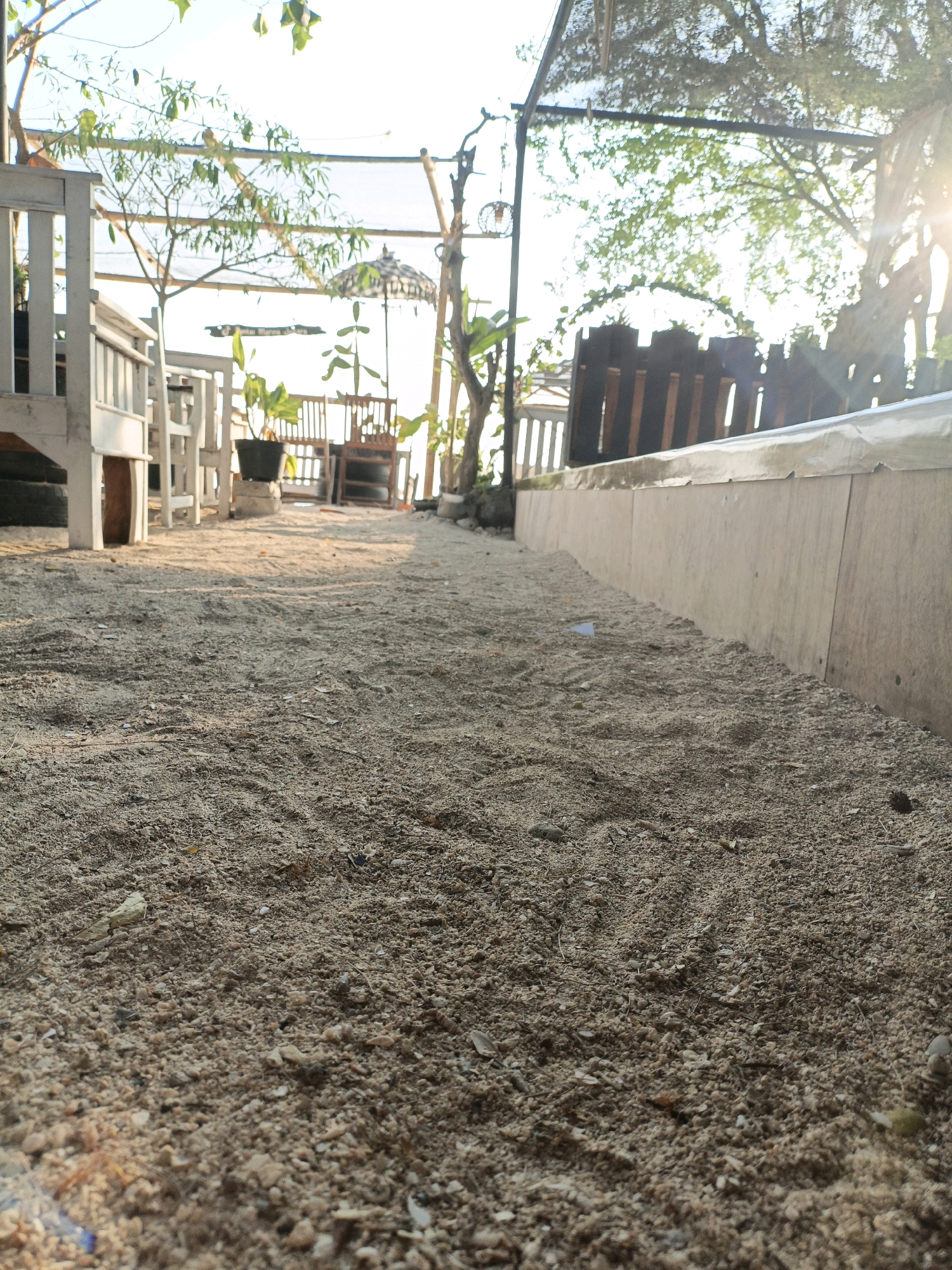 The image depicts a sandy pathway leading towards a beachside seating area. The scene is outdoors, likely in a tropical environment, given the presence of lush greenery and clear, bright sky. The pathway is covered in light brown sand with visible small pebbles and shell fragments. To the left of the pathway, there are white wooden tables and chairs, suggesting a cafe or restaurant setting. Further in the background, more wooden furniture and potted plants are visible, along with a distinctive black and white patterned umbrella. A wooden fence or partition runs along the right side of the pathway, constructed of vertical wooden planks. Beyond the fence, trees and vegetation are present, with sunlight filtering through the leaves, creating a dappled effect. The overall ambiance suggests a relaxed, possibly vacation-oriented location. The description of "Cikal, Indonesia" is not visually verifiable from the image alone.