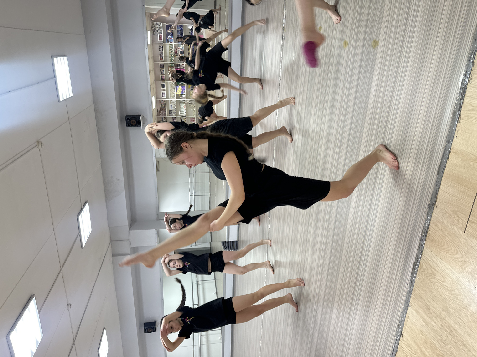 The image depicts a group of girls in a dance studio, likely located in a building in Votkinsk, Russia. The girls, dressed in black shorts and t-shirts, are barefoot and performing a dance routine. They are arranged in rows, with each girl lunging with one leg extended forward and the other leg stretched behind. Their arms are raised above their heads, with their hands touching. The studio has a wooden floor with a grey striped area in the middle. A wall is lined with mirrors, where reflections of the dancers and decorations are visible. The ceiling is white, illuminated by fluorescent lights. A black speaker is mounted on the wall.