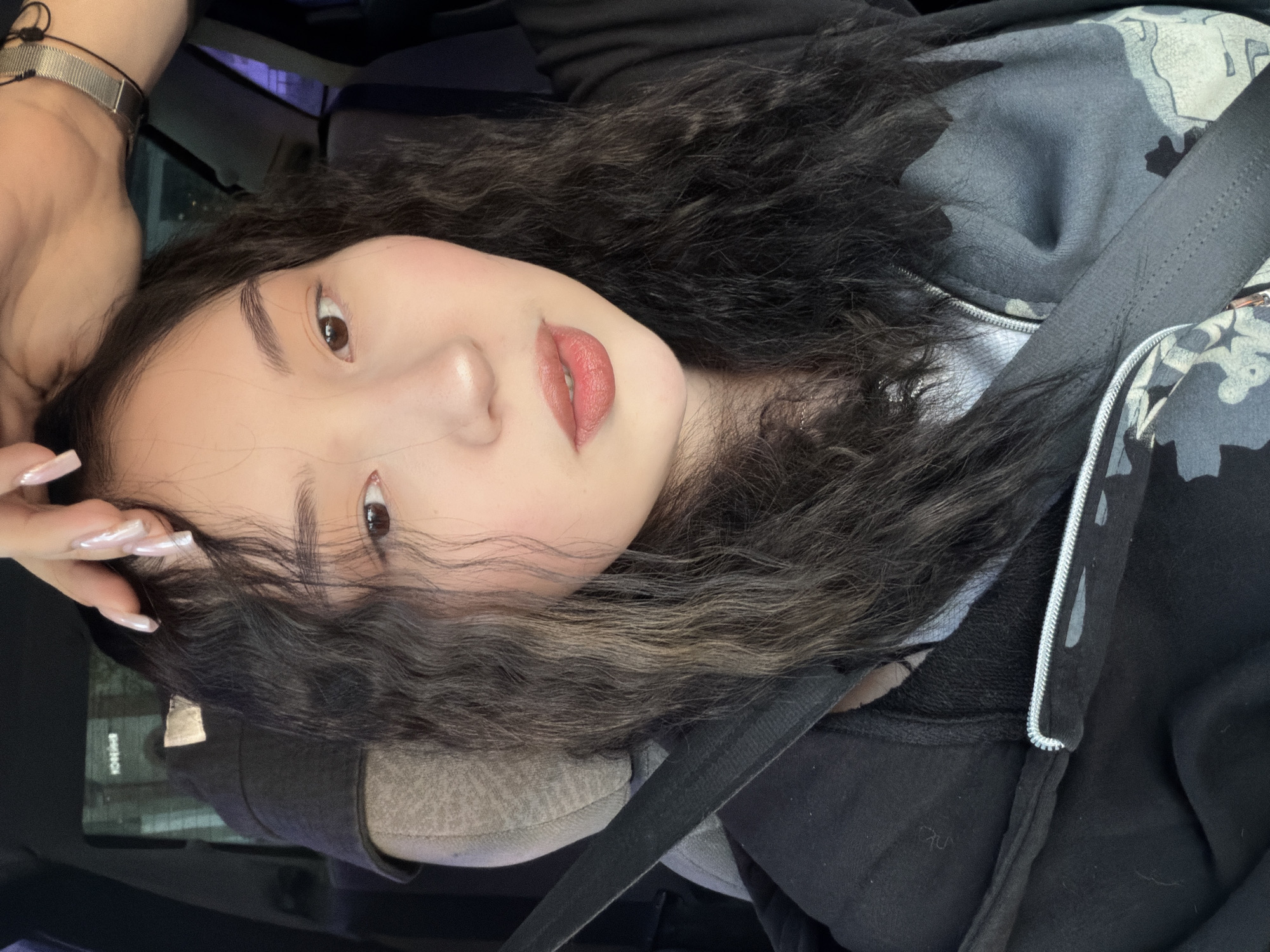 A young East Asian female, with long, dark, wavy hair and fair complexion, is seated in what appears to be the passenger seat of a vehicle, in the city of Chaikovsky, Russia. Her dark brown eyes are directed towards the camera. She has light makeup, including subtle blush on her cheeks and peachy-pink lipstick. Her right hand, adorned with long, light-colored manicured nails, rests near her temple, touching her hair. A silver-colored metal link watch is visible on her left wrist. She is wearing a dark, possibly black, zip-up hooded jacket or sweatshirt, with a silver zipper. Beneath the jacket, a grey and camouflage patterned garment is visible. A dark seatbelt diagonally crosses her chest and shoulder, secured. The car interior features a grey/tan fabric seat and a dark headrest. Through the car window in the background, a blurred, indistinct outdoor scene with faint lights suggests low-light conditions, possibly dusk or night. A faint, inverted reflection of text, possibly "KONAMI", is visible on the window in the upper left.
