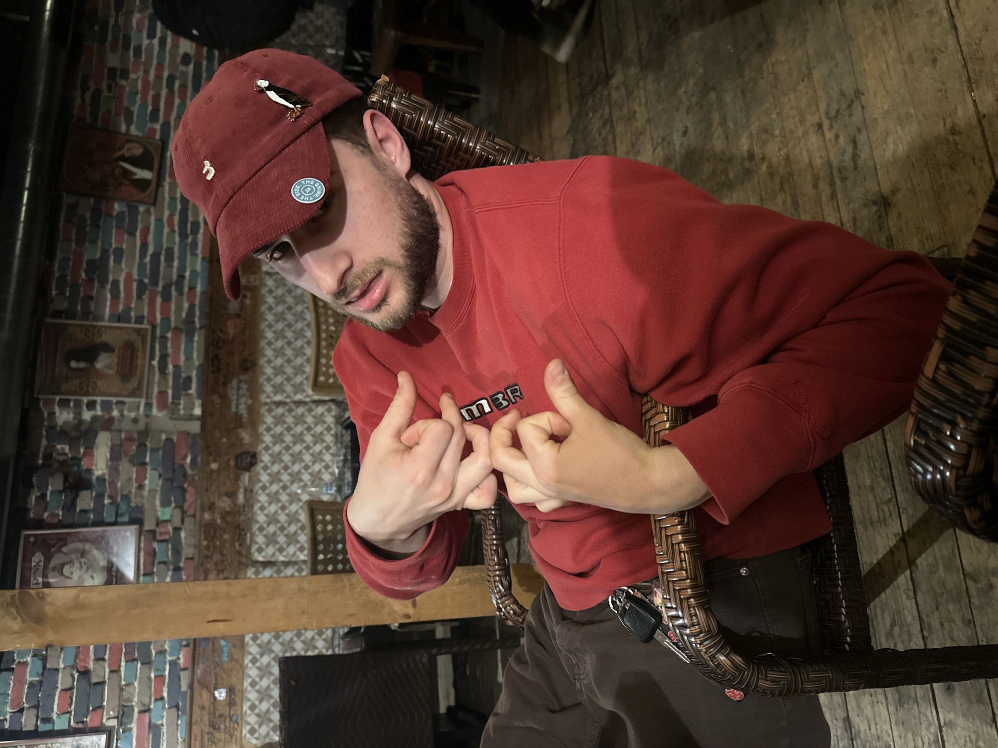 The image depicts a young adult male seated in a dark brown wicker chair within an indoor establishment in the city of Plovdiv, Bulgaria. The subject, positioned centrally, wears a red corduroy baseball cap adorned with a circular "WE RISE & SHINE" pin and a black, white, and gold bird-shaped pin. He has a beard or stubble and wears a red crewneck sweatshirt with the letters "MBR" partially visible on the chest, and dark brown trousers. His hands are interlocked or performing a specific gesture at chest height, with thumbs and index fingers of both hands touching, forming a shape. A set of black car keys is clipped to his left trouser pocket. He leans back, looking towards the right with his mouth slightly ajar. The background features a distressed brick wall composed of red, blue, grey, and black bricks, on which several framed artworks are hung, including a portrait, an image with "PNK" text and a figure, and a depiction of a person playing a saxophone. A vertical wooden beam is visible behind the subject. The flooring consists of dark, worn wooden planks. In the far right background, a partial view of another individual's leg, clad in dark trousers and a sneaker, is visible. The lighting is dim, suggesting an evening or interior setting.