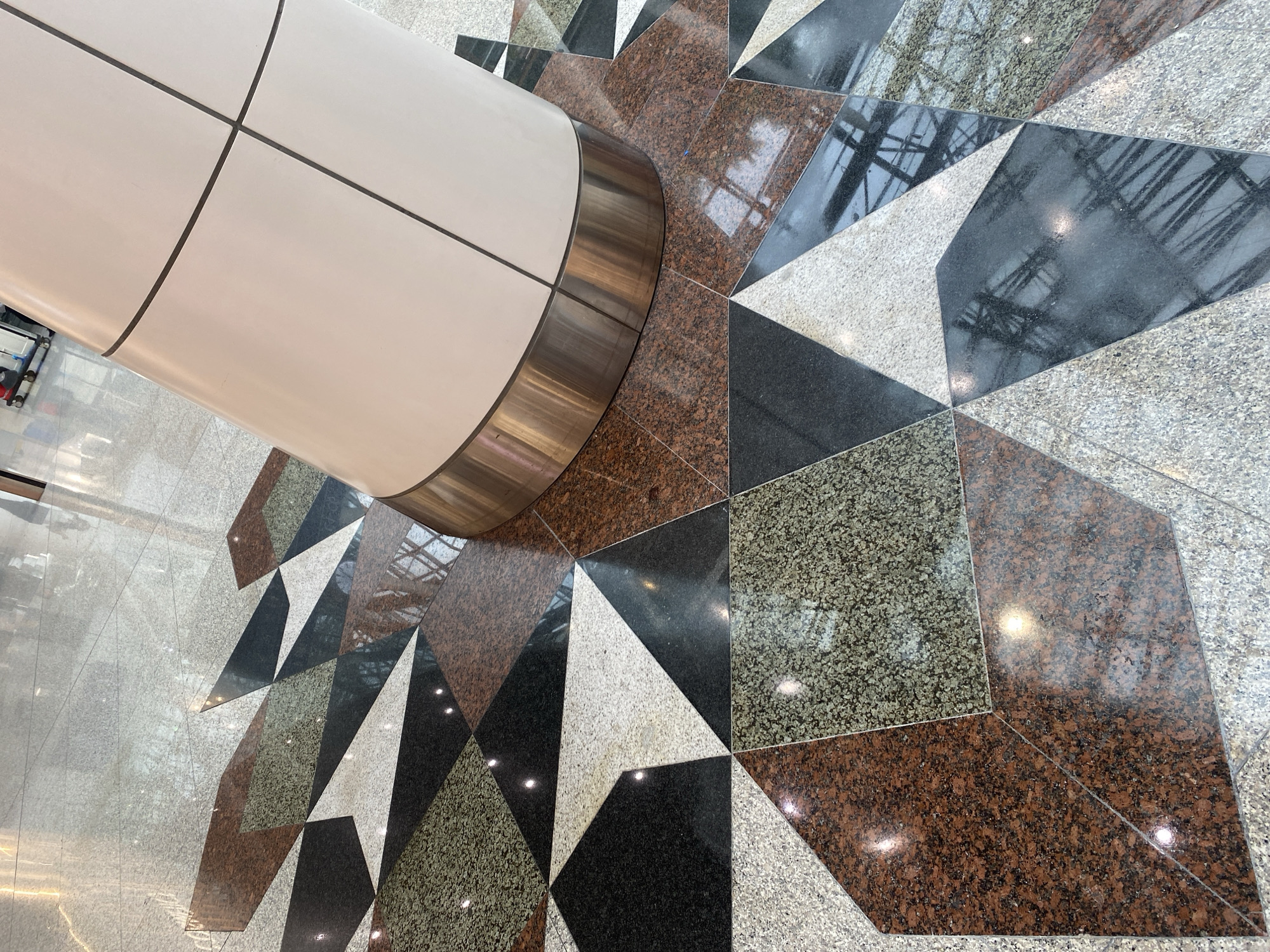 The image shows a close-up view of a polished granite floor in a modern building. The floor is laid with geometrically shaped tiles in various colors including black, white, speckled gray, and reddish-brown. A large, cylindrical white pillar with metallic trim stands in the center of the frame. Reflections of lights and architectural elements are visible on the shiny surfaces of the floor and pillar. The overall impression is one of a clean, well-maintained indoor space, likely a public area like an airport terminal or a shopping mall. Based on the visual cues, this scene could be situated within KLIA (Kuala Lumpur International Airport) in Sepang, Malaysia, a prominent example of modern Malaysian architecture featuring such intricate flooring designs.