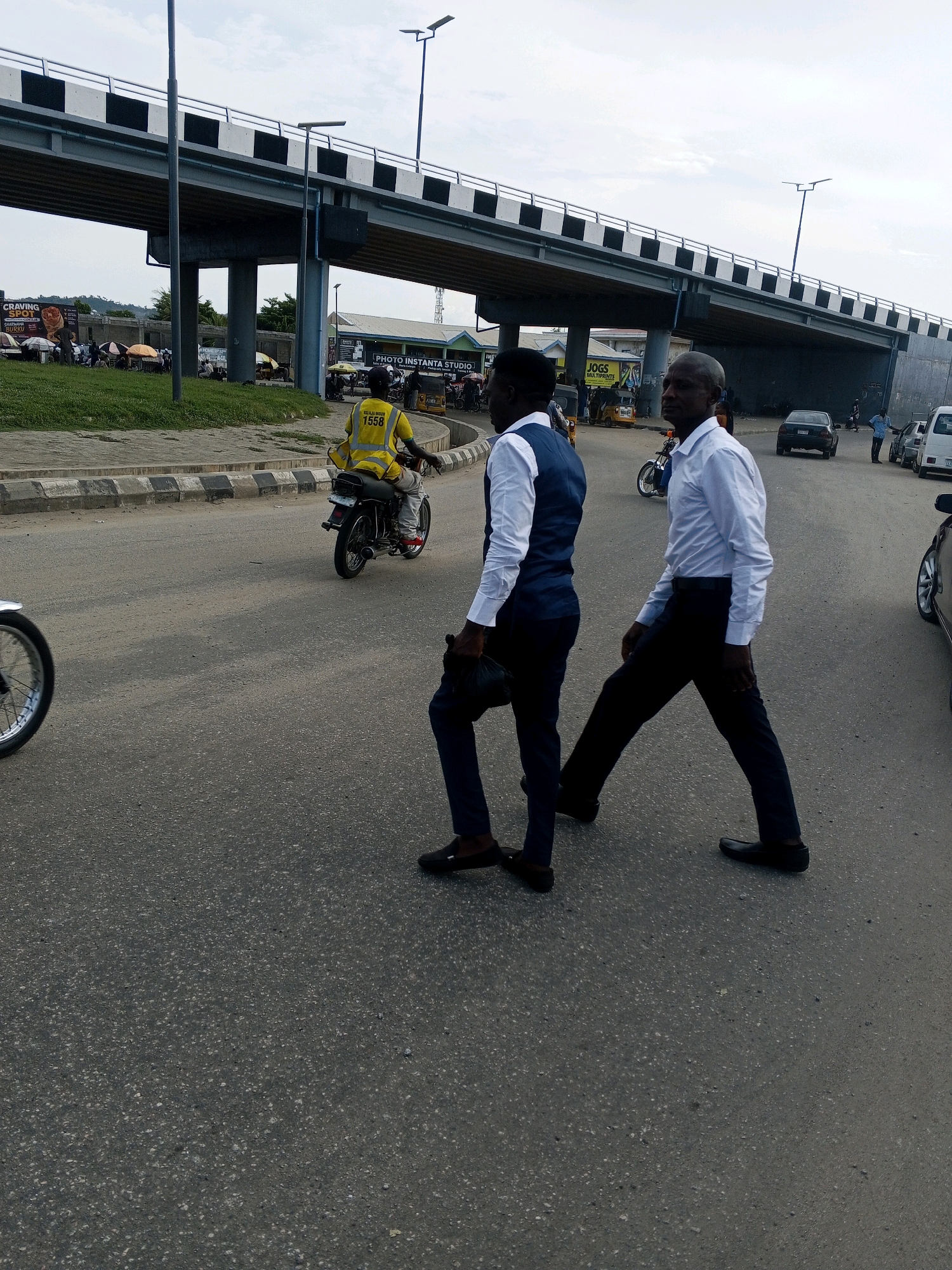 The image depicts a street scene in Lokoja, Nigeria, beneath an elevated highway overpass. Two men in formal attire are walking across the asphalt road. The man on the left is wearing a white shirt and a navy blue vest, with dark trousers and dark shoes, carrying a black bag in his left hand. The man on the right is wearing a white long-sleeved shirt and dark trousers, with dark shoes. A motorcycle rider in a bright yellow vest with the number "1558" is visible in the background, moving away from the viewer. Several other vehicles, including cars and a white van, are also present on the road. The environment includes a grassy area to the left of the road, lined with a concrete curb. In the distance, there are various shops and stalls, with umbrellas providing shade. The sky is overcast.