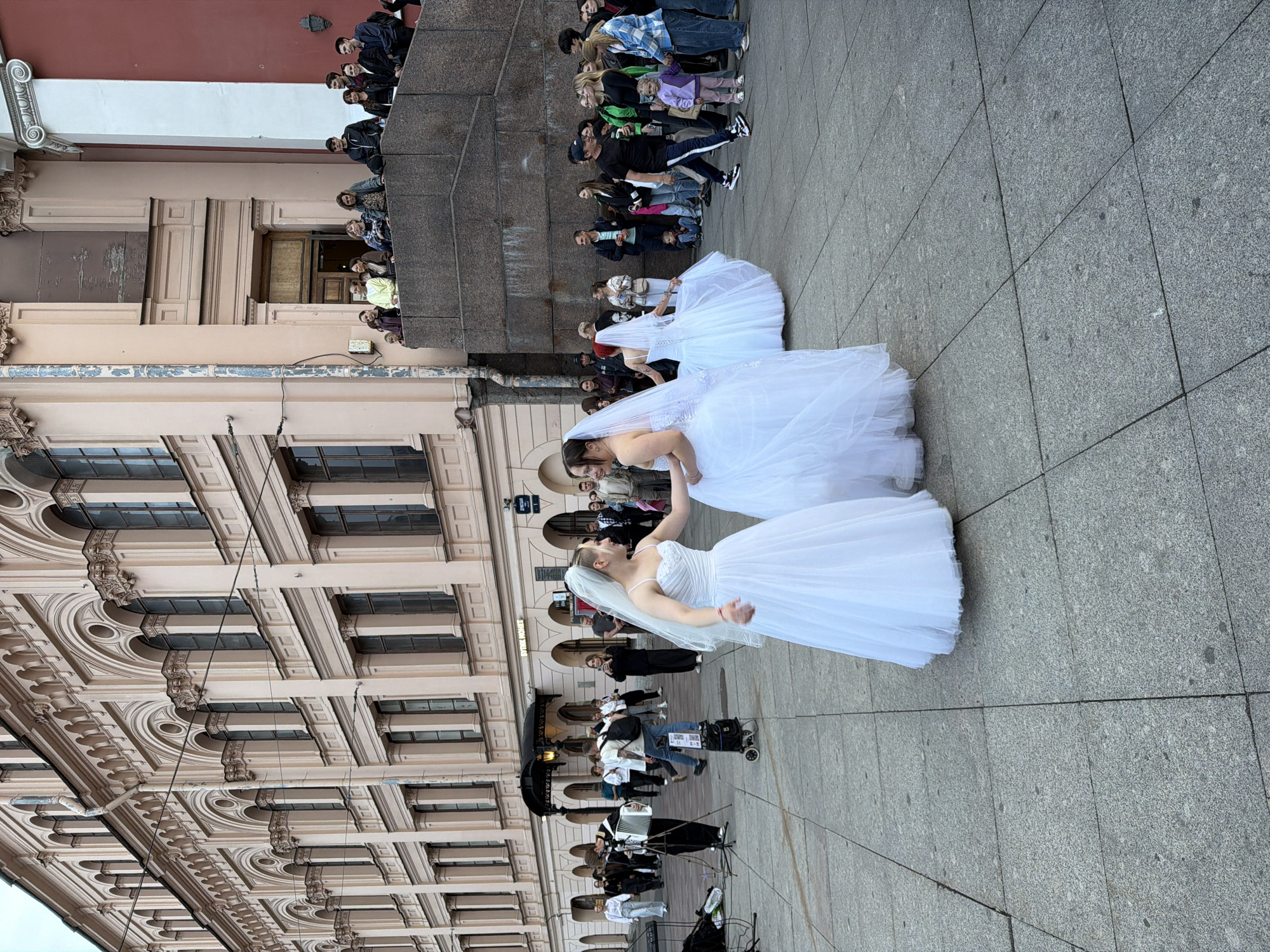 In Saint Petersburg, Russia, on a grey, paved plaza in front of a large, ornate building with cream-colored facades and arched windows, three women in white wedding dresses are dancing. The woman on the left, with short hair and a veil, is facing right, her arms outstretched. To her right, another woman in a wedding dress is dancing, her back to the viewer, her veil trailing. A third woman in a wedding dress, partially visible, is to the right of the second woman.
A crowd of onlookers is gathered on a raised stone platform behind the dancers, observing the scene. To the left of the dancers, a person plays an accordion, and a keyboard and speaker are set up on the plaza. The building in the background features multiple stories with repeating architectural elements, including arched windows and decorative cornices. The ground is made of large, square grey paving stones.