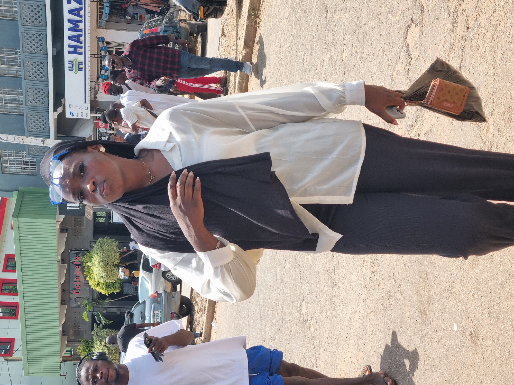 The image depicts a street scene in Jos, Nigeria. The primary subject is a woman in the foreground, dressed in a white blouse and black pants, with a black scarf draped over her shoulders. She is looking towards the camera, with a neutral expression. She is holding a black plastic bag containing a brown patterned wallet. Beside her, to the left, a man wearing blue shorts and a white t-shirt is partially visible. He is holding a phone and wearing headphones.
In the background, other people are visible, some walking and others standing near vehicles and buildings. One man in a red and black checkered shirt is prominent. A white building with the text "HAMAZ" in blue and green lettering is visible, along with a green building with red-trimmed windows and text that appears to be related to a restaurant. Cars are parked on the street. The lighting suggests it is daytime, likely late afternoon due to the long shadows cast on the ground. The weather appears to be clear. The overall impression is of a busy, everyday street scene.