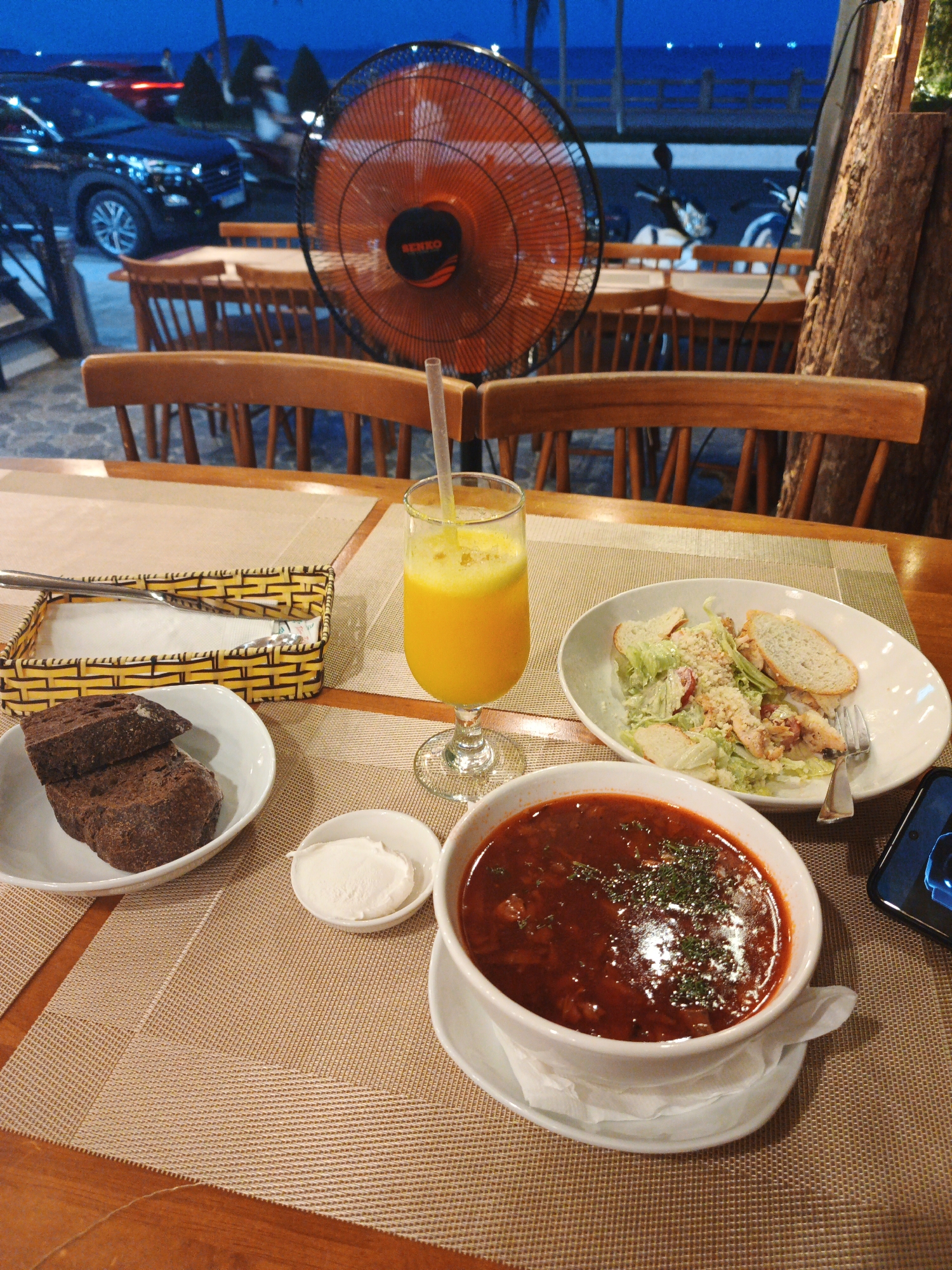 The image depicts an outdoor dining scene at dusk, likely in Bac Nha Trang Ward, Vietnam. A table is set with a meal consisting of a bowl of hearty red soup, possibly borscht, garnished with herbs; a salad with greens, croutons, and what appears to be chicken; two slices of dark bread in a white bowl; and a small dish of what looks like sour cream or cheese. A glass of bright orange juice with a straw sits next to a woven basket containing napkins and cutlery.
In the background, wooden chairs and tables are arranged, suggesting a restaurant or cafe setting. A large, orange industrial fan is prominently placed, indicating a warm climate. Beyond the dining area, a street scene is visible with cars and motorbikes. The sky is a deep blue, transitioning into twilight, with a glimpse of a body of water and lights in the distance. The overall atmosphere seems relaxed and casual, with the setting suggesting a pleasant evening meal by the coast. No people are clearly visible in the foreground, though indistinct figures can be seen in the background. The visible text on the fan reads "RENKO".