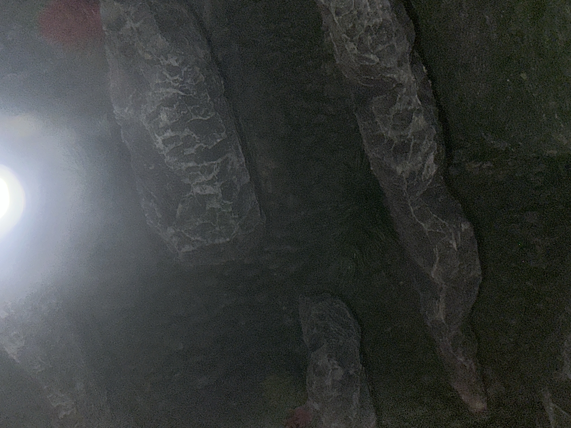 The scene depicts a dimly lit, interior, subterranean environment. A single, intense, circular white light source is visible at the top-center, emitting a strong glare and illuminating a localized hazy area. The predominant surfaces are dark, appearing as architectural cladding or structural elements. These surfaces display intricate, irregular, lighter-colored patterns that resemble natural stone veining or abstract topographical reliefs. These patterns are visible across multiple distinct sections of the dark surfaces within the frame. No identifiable individuals, distinct objects, or specific activities are present. The architectural styling and material characteristics are consistent with the grand underground infrastructure of the Moscow Metro system, located in Moscow, Russia.
