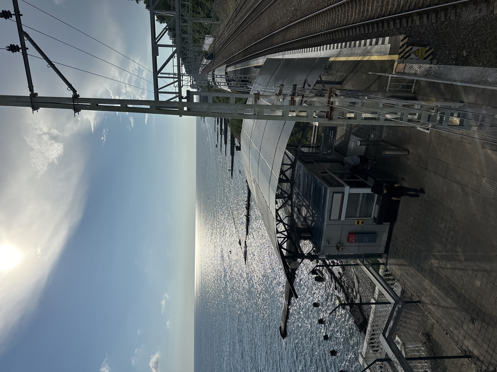 The image depicts a train station platform situated on the coast of Sochi, Russia. The sun is shining brightly, casting a strong reflection on the calm sea. The platform features a modern, covered waiting area with a translucent roof. Adjacent to the platform are the train tracks, with a train visible in the distance. A tall metal structure supporting overhead power lines stands prominently on the right side of the frame. The environment includes a rocky coastline with some breakwaters. Several people are present on the platform, including individuals walking and standing. The scene appears to be during daylight hours, likely in the late afternoon given the angle of the sun.