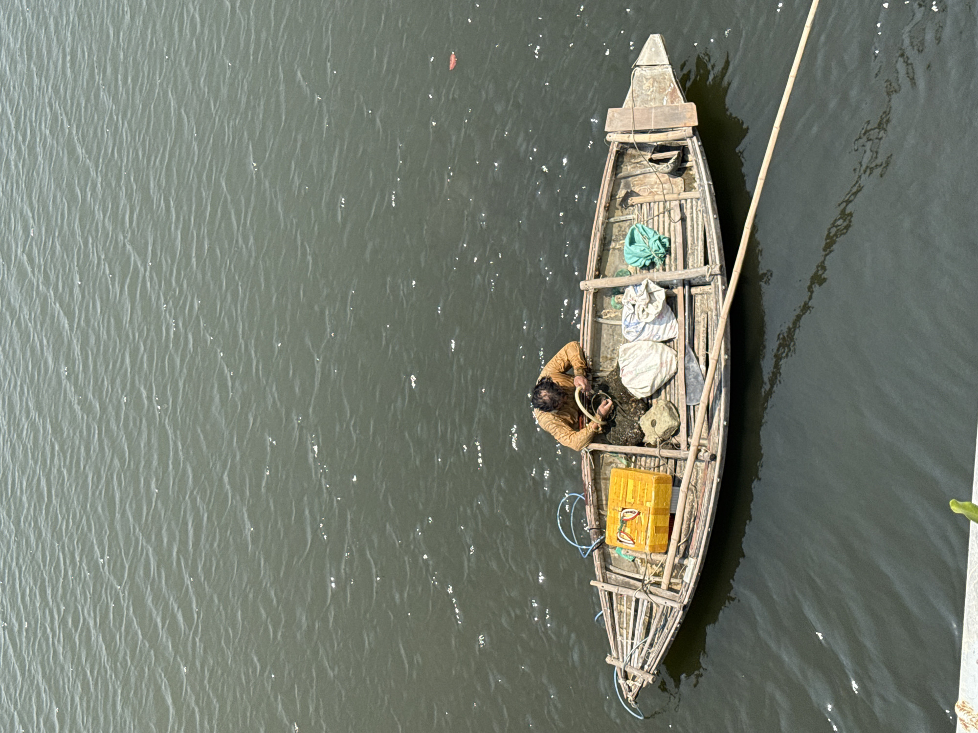 Captured from an elevated perspective, the image shows a narrow, wooden boat positioned on a body of water. Inside the boat, an individual is engaged in an activity that seems related to fishing, managing rope and nets. The boat contains various items, including a yellow box, bags, and other fishing-related objects. The water surrounding the boat displays a subtle texture and slight ripples on its surface. The lighting suggests it is daytime. The location appears to be Đà Nẵng, Vietnam, based on the image's visual characteristics.