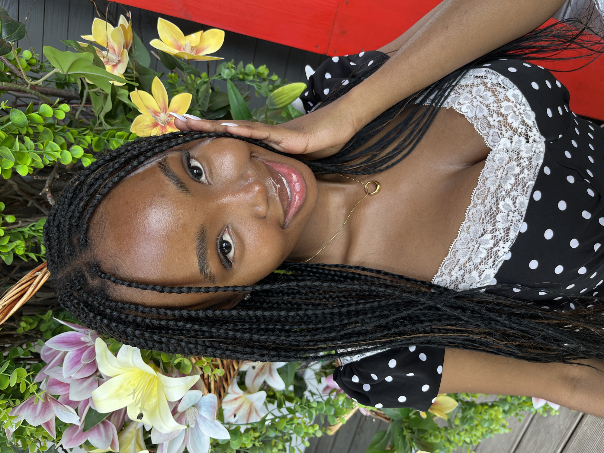 The image shows a close-up of a young Black woman with braided hair, wearing a black and white polka dot dress with a white lace trim. She is smiling and touching her face with her right hand. She is wearing a gold necklace with a circular pendant. She is positioned in front of a backdrop of artificial flowers and greenery, with a red object visible on the right side of the frame. The setting appears to be outdoors, possibly a decorative display. There is no indication of the location being in Kazan, Russia.