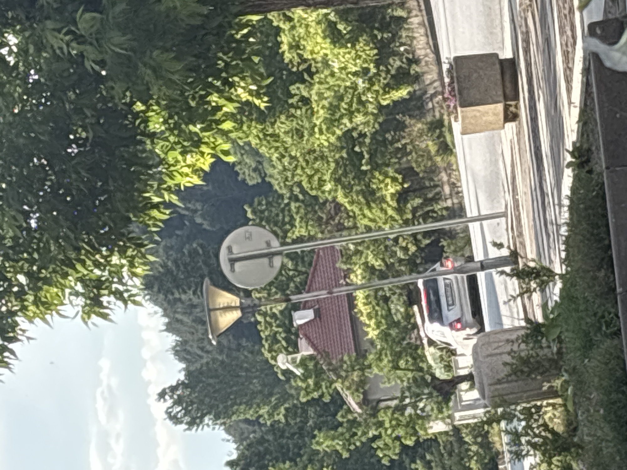 A sideways view captures a street scene in Garmen, Bulgaria. The road is lined with trees and vegetation. A traffic sign indicating a pedestrian crossing is prominent. A car is visible on the street. Buildings with red roofs are also visible. The sky is partly visible between the foliage. Stone objects are on the roadside.