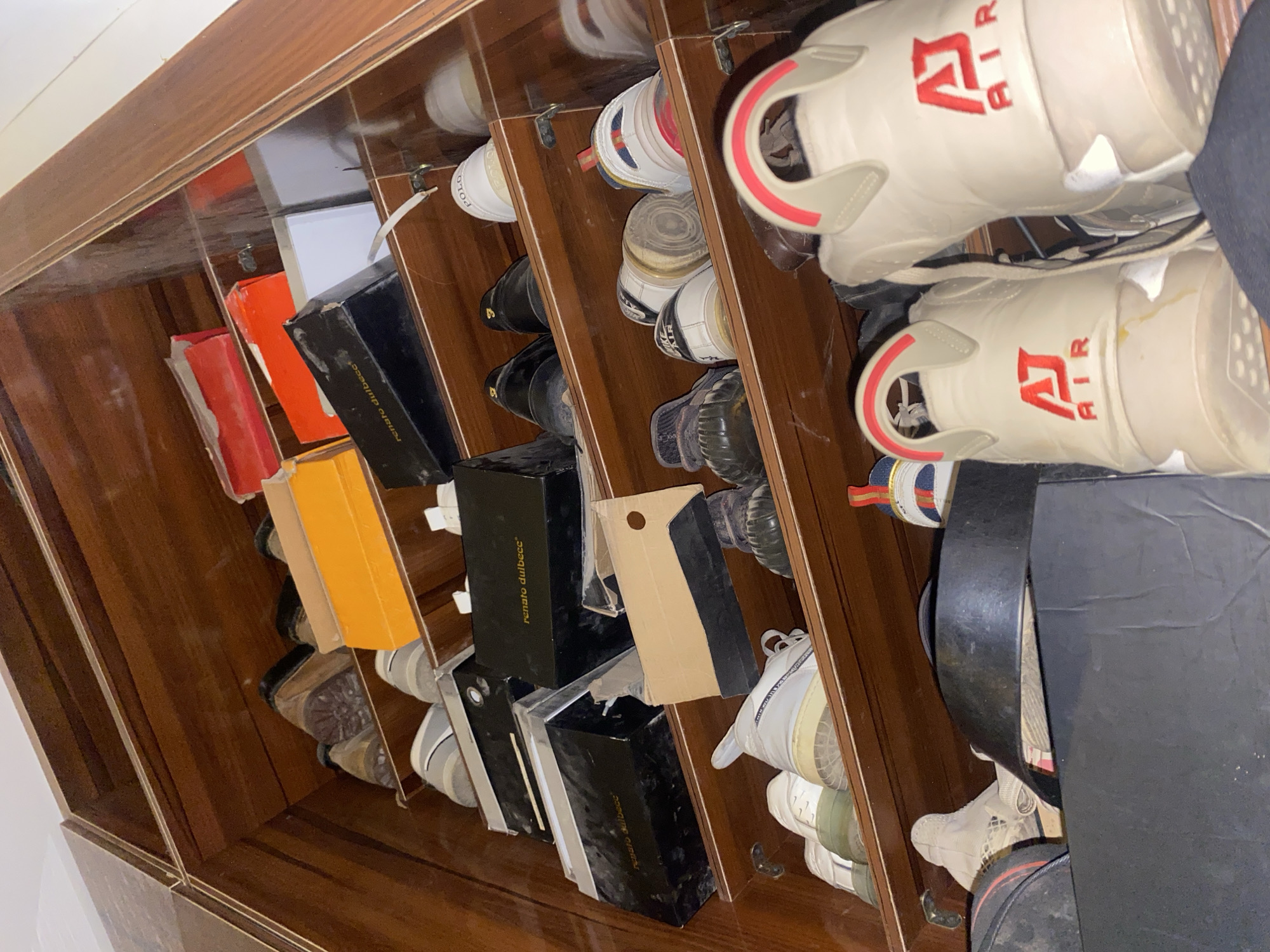 The image depicts a shelf unit filled with various footwear, primarily sneakers and some dress shoes, arranged on wooden shelves. The items are a mix of new and used, with some still in their original boxes. On the top shelf, there are red and orange shoeboxes, alongside dark-colored shoes. The middle shelves contain more shoeboxes, some black with gold lettering that appears to read "renato dulbeccce," as well as individual shoes. The bottom shelf shows a collection of sneakers, some with prominent branding like "POLO," "NIKE AIR," and what appears to be "AJ AIR." The overall environment appears to be an indoor storage space, possibly a closet or a dedicated shoe storage unit. The lighting is somewhat dim, suggesting artificial light sources. No people or specific geographical markers of Giginyu, Nigeria are visible in the image.