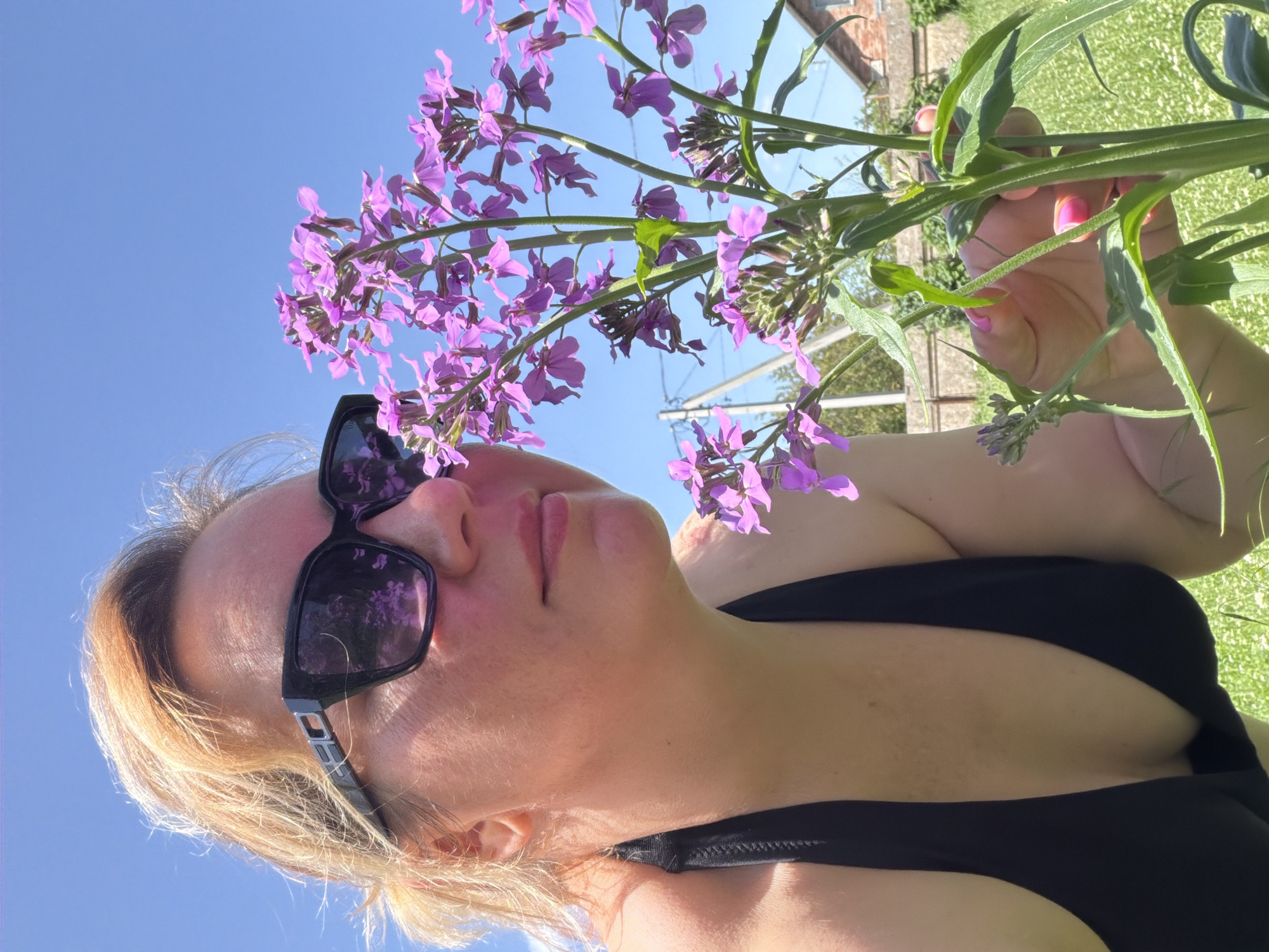 A woman wearing sunglasses and a black garment is holding a flowering plant with purple blossoms. The sky is blue. The location is Leningradskaya, Russia.