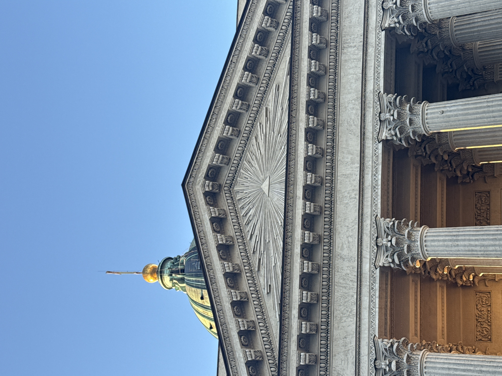 This is a low-angle shot of a section of the Kazan Cathedral in Saint Petersburg, Russia. The image primarily showcases the neoclassical architectural elements of the building. On the right, fluted columns with Corinthian capitals are visible, arranged in a tiered manner. Adjacent to the columns is a wall adorned with decorative molding and small, repeating square or rectangular motifs. Moving towards the left, there is a triangular pediment featuring a radiating sunburst design in the center, surrounded by layers of ornamental details. In the top-left of the image, a portion of the cathedral's dome is visible against a clear blue sky. The dome is green, topped with a golden spherical element and a spire. The overall lighting suggests a sunny day.