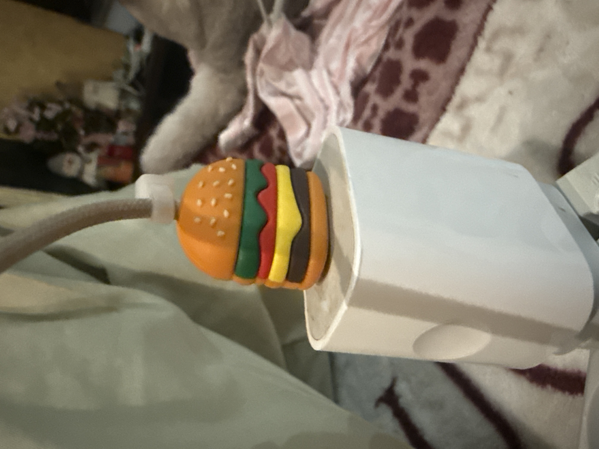 A white power adapter is plugged into an electrical outlet. Attached to the cable, near the adapter, is a small, colorful charm shaped like a hamburger. The charm has layers representing a bun, lettuce, tomato, cheese, and patty, with the bun being orange and dotted with seeds. The cable itself is gray and appears to be flexible. The background is blurred and suggests an indoor setting with soft furnishings, possibly a bed or sofa with patterned blankets. A light-colored object, possibly a piece of fabric or another person's limb, is partially visible in the upper left of the frame. The lighting is artificial and the image appears to have been taken at close range. There is no visible text in the image.