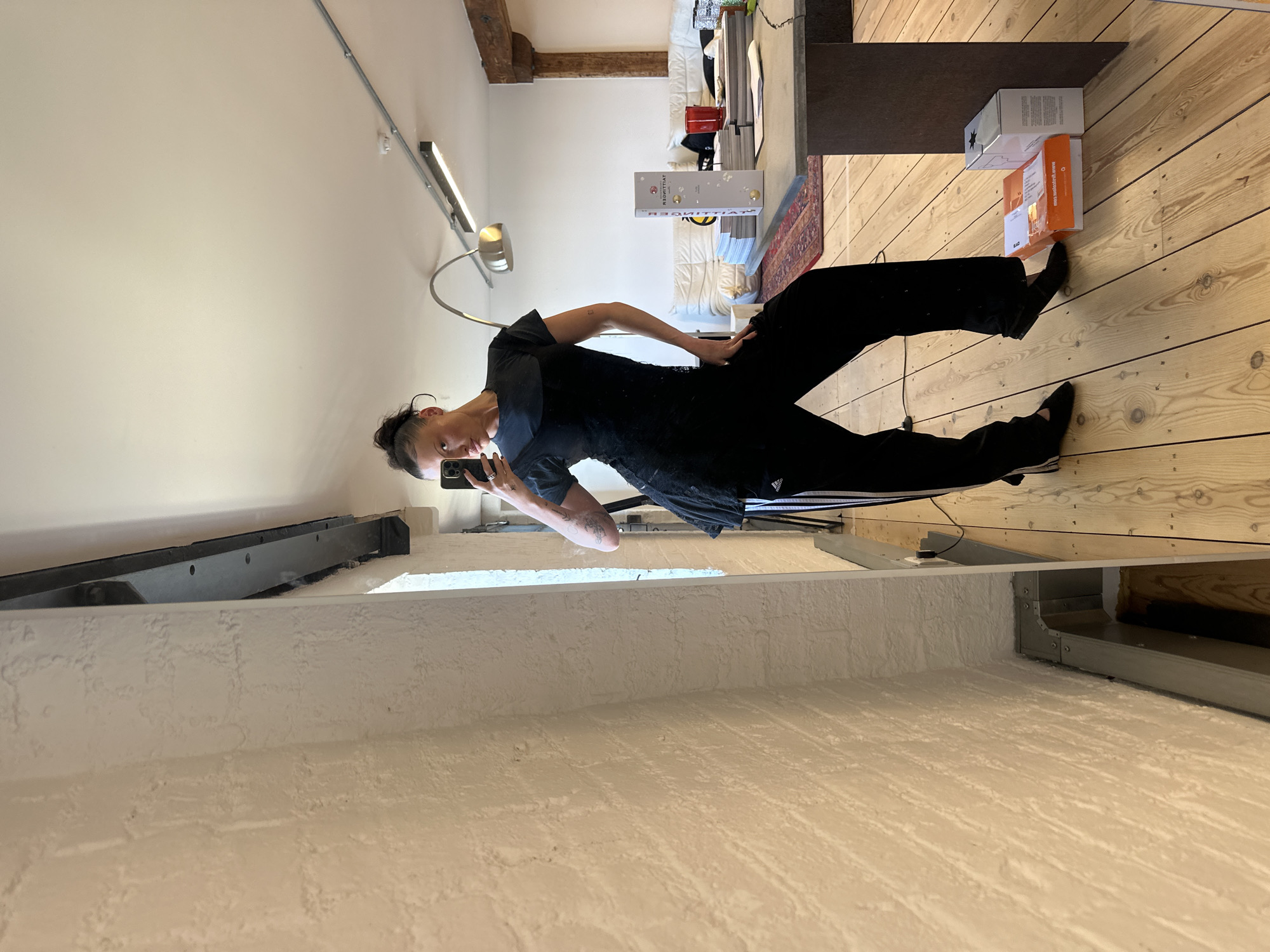 A woman stands in front of a mirror in an indoor setting, possibly an apartment or loft. She is holding a smartphone and taking a selfie, wearing a black t-shirt and black pants with white stripes down the side. The environment suggests a living space with hardwood floors, a white wall with a visible structural beam, and a desk or shelf with various objects including boxes and books. Natural light filters into the room. The scene lacks any defining characteristics that would specifically place it in London, United Kingdom.