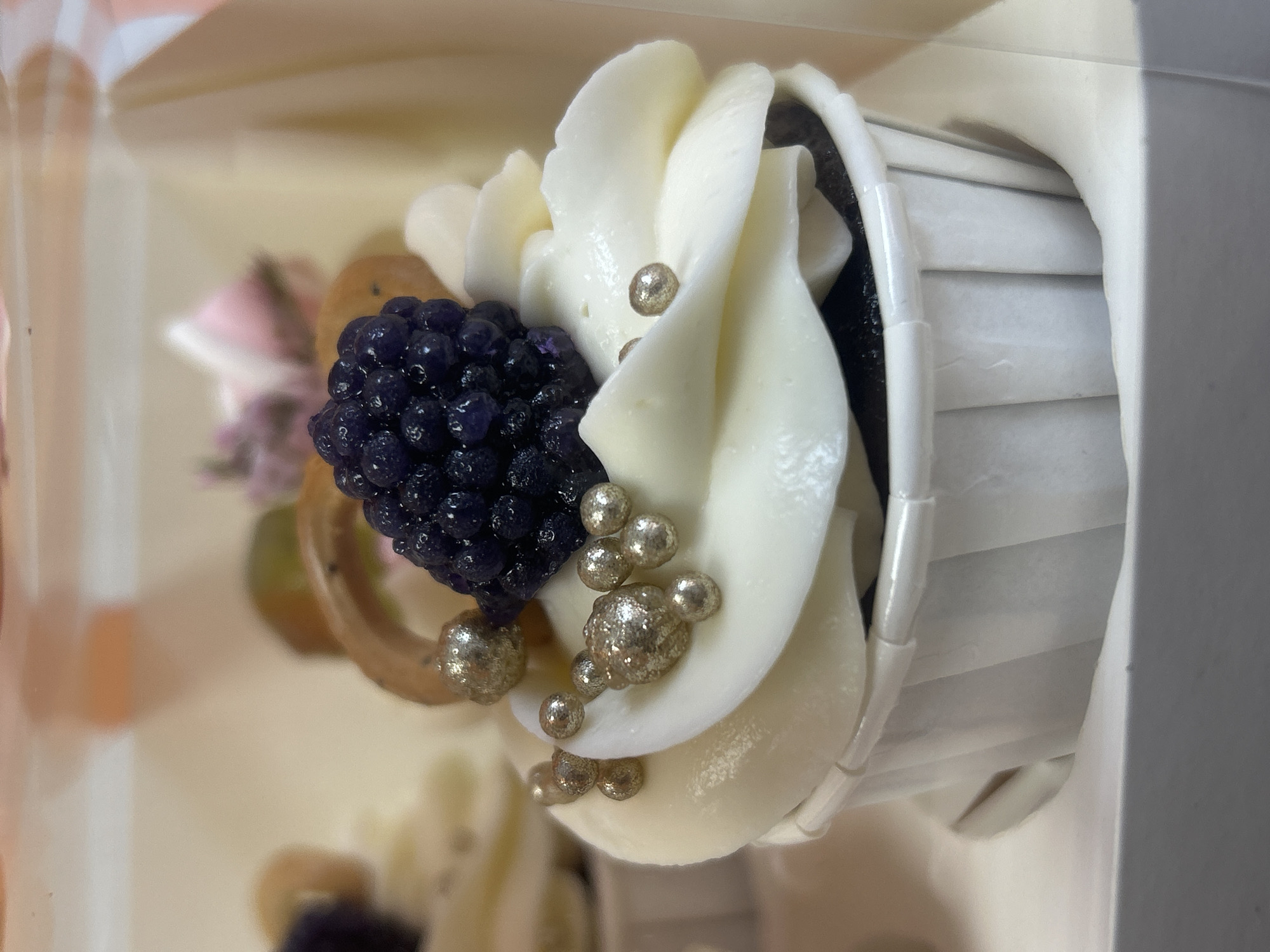 The image shows a cupcake in a white paper cup. It is decorated with white frosting, a cluster of dark purple berries, and small gold-colored balls. There is another cupcake visible behind this one. The image appears to be taken in a container, such as a bakery box. The location is not evident from the image.