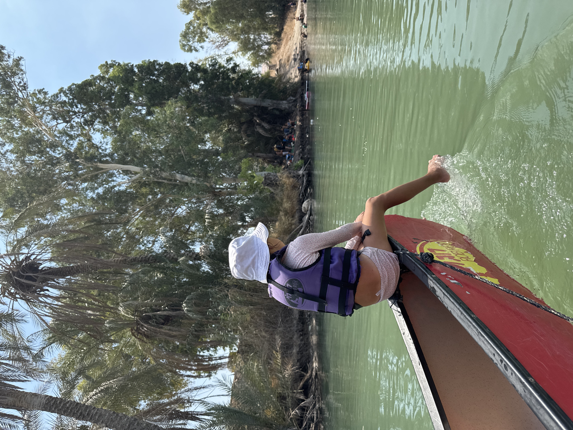 In Emek HaYarden Regional Council, Israel, a young girl sits on the edge of a red canoe, her legs dangling in the emerald green water. She wears a white bucket hat and a purple life vest over a light-colored rash guard and patterned shorts. Her right foot is submerged in the water, creating ripples and splashes. The canoe is partially visible in the foreground, with a yellow logo on its side. The background features a lush green riverbank lined with palm trees and other dense foliage. In the distance, a sandy bank is populated by several people, some sitting and others standing, with various inflatable watercraft scattered on the water. The sky above is clear and blue.