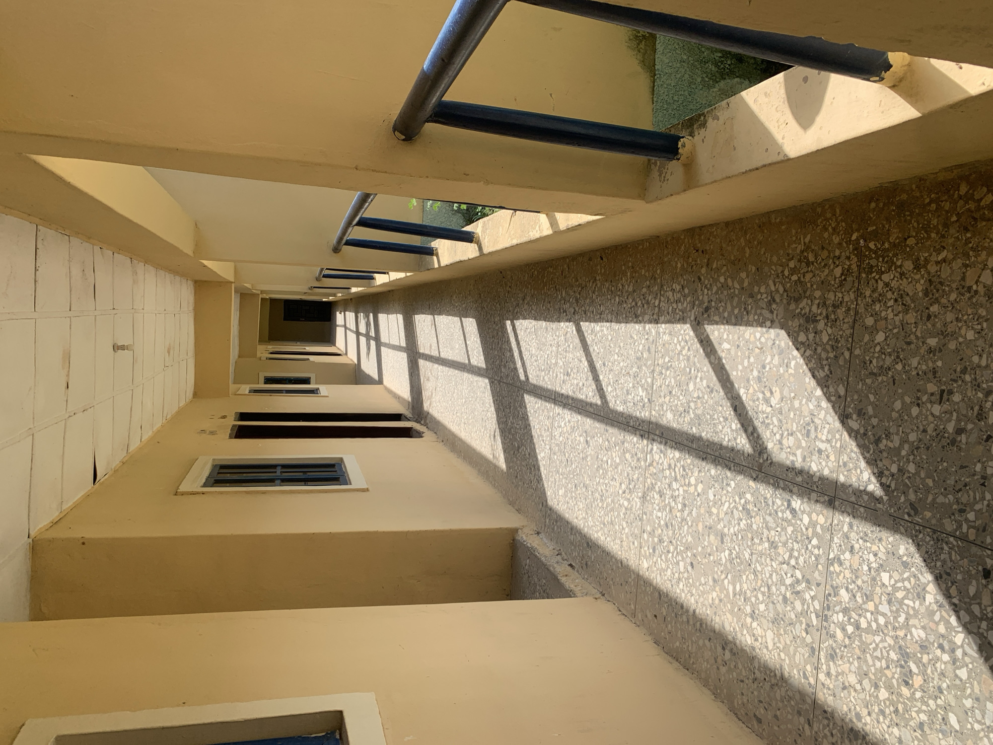 The image depicts a long, exterior hallway of a building, likely in Jalingo, Nigeria. The hallway has a pale yellow-painted exterior wall on the left, featuring several windows with blue frames. On the right side of the hallway, there's a railing with dark blue vertical supports and a metal handrail. Sunlight streams in from the right, casting long, geometric shadows across the speckled, grey-and-white tiled floor. The ceiling appears to be made of white tiles, some of which show signs of wear. The hallway extends into the distance, with doorways visible on the left side. The overall impression is of a simple, functional outdoor corridor in a warm, sunny climate.