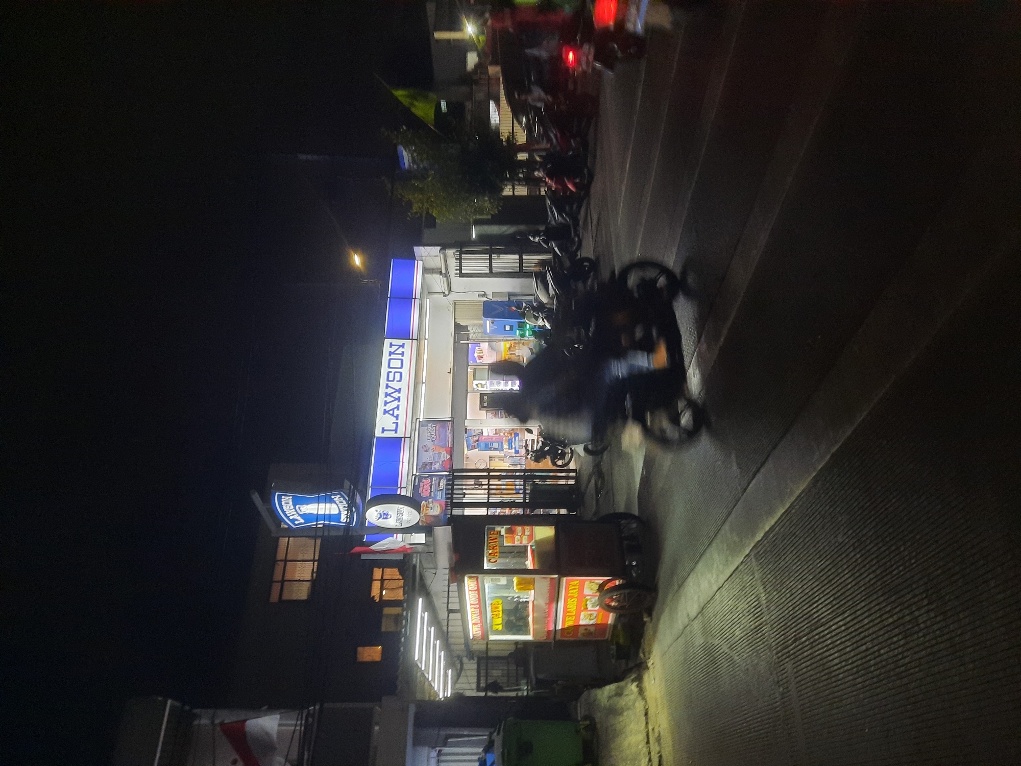 At night, a food cart is positioned on the left side of the image, in front of a brightly lit Lawson convenience store in North Jakarta, Indonesia. The food cart, which appears to sell fried food like "Cakwe," has signs with Indonesian text. Behind the food cart, the Lawson store has a blue and white illuminated sign and its interior is visible, showing shelves stocked with products and a cashier area. Several motorcycles are parked in front of the Lawson store and on the street. A person on a motorcycle is in motion in the foreground, blurring the image slightly. The street pavement has distinct textured lines. The scene is illuminated by the store's lights and a street lamp.