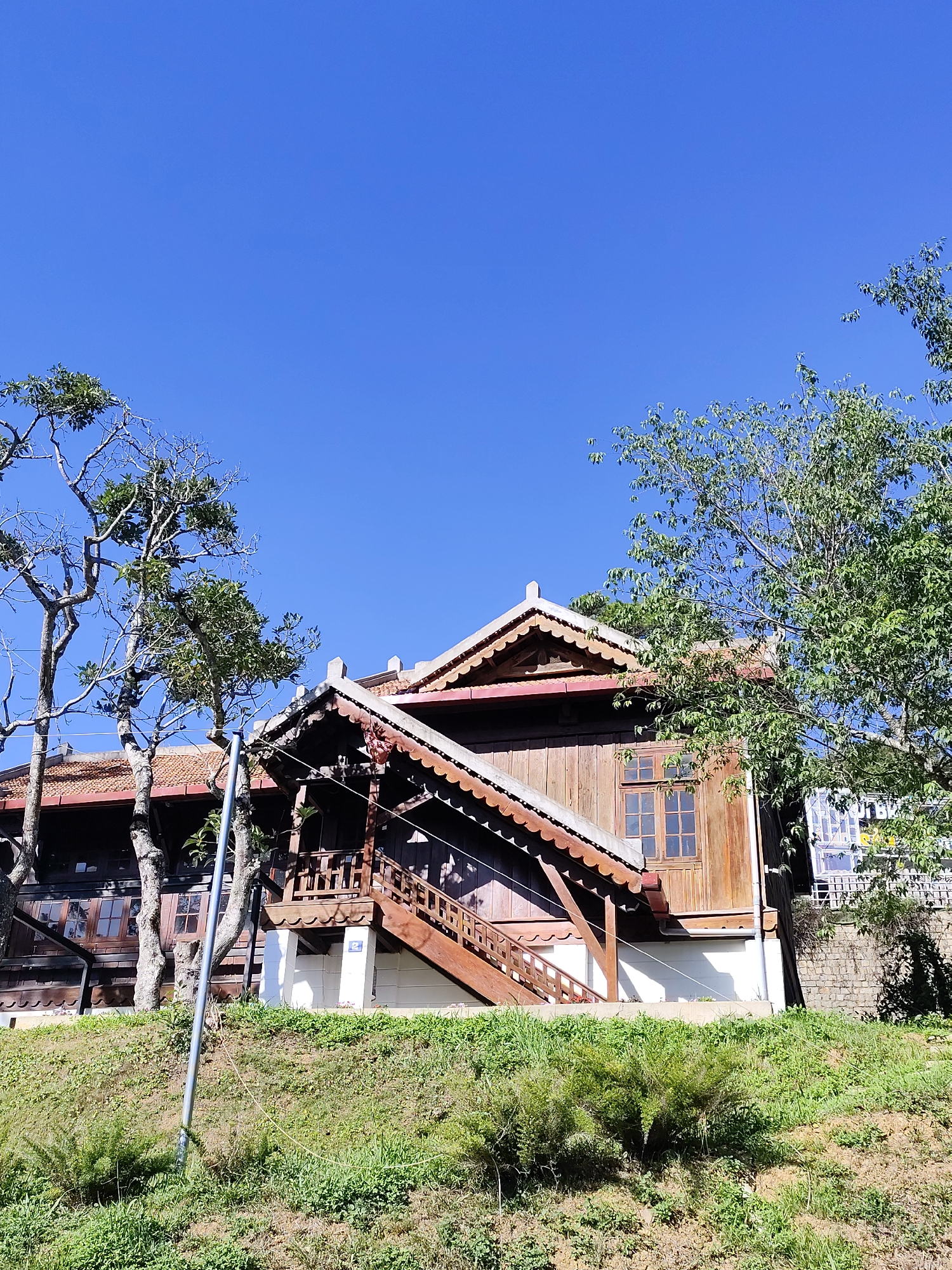 The image captures a distinctive wooden structure nestled in the city of Da Lat, Vietnam. The building appears to be a two-story wooden house featuring traditional Vietnamese architectural elements. It has a steeply pitched roof with scalloped edges, indicative of local design. The wooden facade is weathered and painted in shades of brown. A set of wooden stairs leads up to a landing, which is supported by white pillars.
The house is situated on a grassy hillside, creating a slight elevation. A few trees, some with sparse foliage, frame the house on either side. The sky overhead is a clear, bright blue.