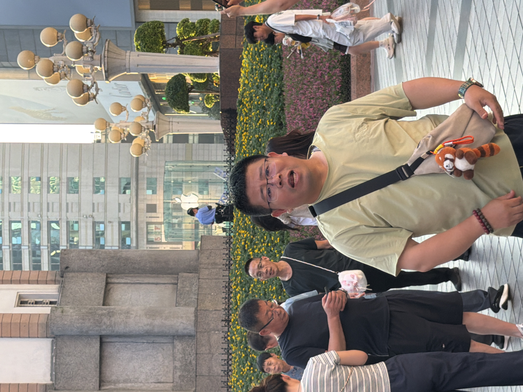 In Yuzhong District, China, a man wearing a light green t-shirt and glasses is in the foreground of the image, facing the camera with his mouth slightly open. He has a beige crossbody bag with a red panda keychain attached, and a beaded bracelet on his left wrist. Behind him, several other people are visible, some walking and others standing. In the background, there are buildings, streetlights adorned with multiple lamps, and a bed of yellow flowers. A person in a blue uniform is also seen in the distance near a building with an illuminated Apple logo. The ground is paved with interlocking tiles.