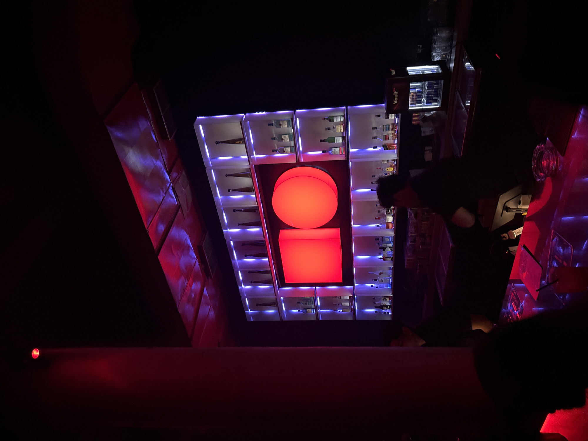 The image captures a dimly lit interior, likely a bar or nightclub in North Kuta, Indonesia, dominated by deep shadows and vibrant artificial lighting. A prominent red-lit column is visible on the left, casting a strong red hue. The background features a well-stocked bar with bottles displayed on illuminated shelves, bathed in a cool purple light. Two large, circular, red-lit panels are centrally positioned on the bar shelves, creating a focal point.
In the foreground, a bar counter is visible, with a few people present, though largely obscured by the low lighting and their dark attire. One person, possibly a bartender, is seen behind the counter, partially illuminated. On the counter, there are various items including what appears to be menus or placemats, an ashtray, and a drink with a straw. A compact, illuminated refrigerator unit with the word "Vault" is also present on the counter. The overall ambiance is one of a modern, atmospheric entertainment venue.