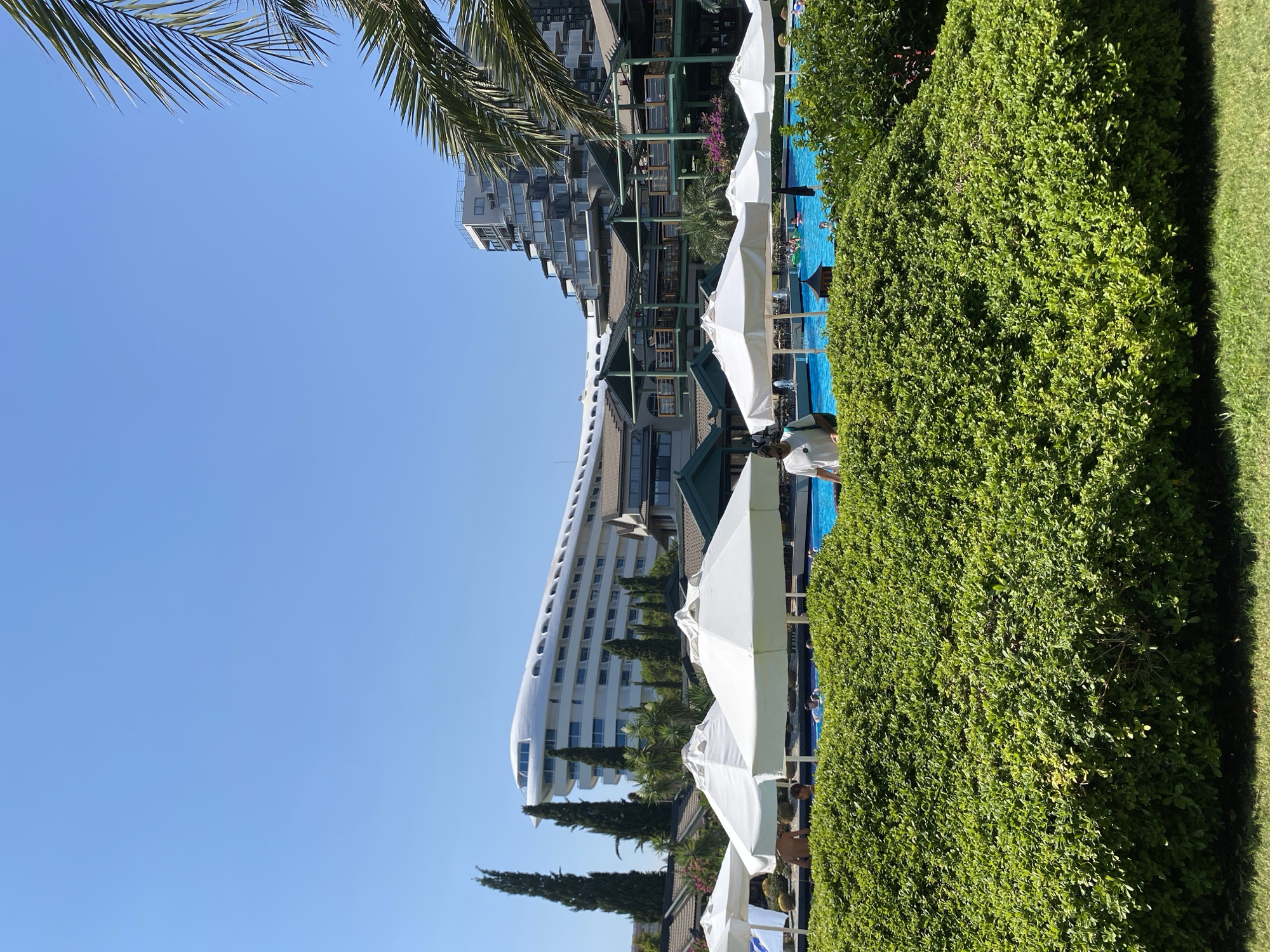 This is an outdoor, eye-level shot of a resort on a sunny day. In the foreground, a meticulously trimmed green hedge runs horizontally across the frame, with a patch of green grass at the bottom left. Behind the hedge, a bright blue swimming pool is visible, with several white umbrellas positioned along its edge. A few people are seen in and around the pool, some in the water and others standing near the umbrellas.
In the mid-ground, there are several multi-story buildings with a resort-like architecture. The buildings feature a mix of dark green roofs, wooden balconies, and cream-colored facades. Some sections of the buildings are stepped or terraced, creating a layered appearance.
Dominating the background is a distinctive, large white building with a curved, wave-like roofline, which appears to be a hotel. This building is positioned to the left of the frame, with its sleek modern design contrasting with the more traditional resort buildings. Tall, slender evergreen trees are scattered throughout the scene, particularly between the foreground and the resort buildings, and to the left of the white hotel. A palm frond is visible in the upper right corner, suggesting a tropical or Mediterranean climate. The sky is a clear, bright blue, indicating a cloudless day.
The location is identified as Aksu, Turkey. The overall scene depicts a vacation resort environment with ample recreational facilities and hotel accommodations.