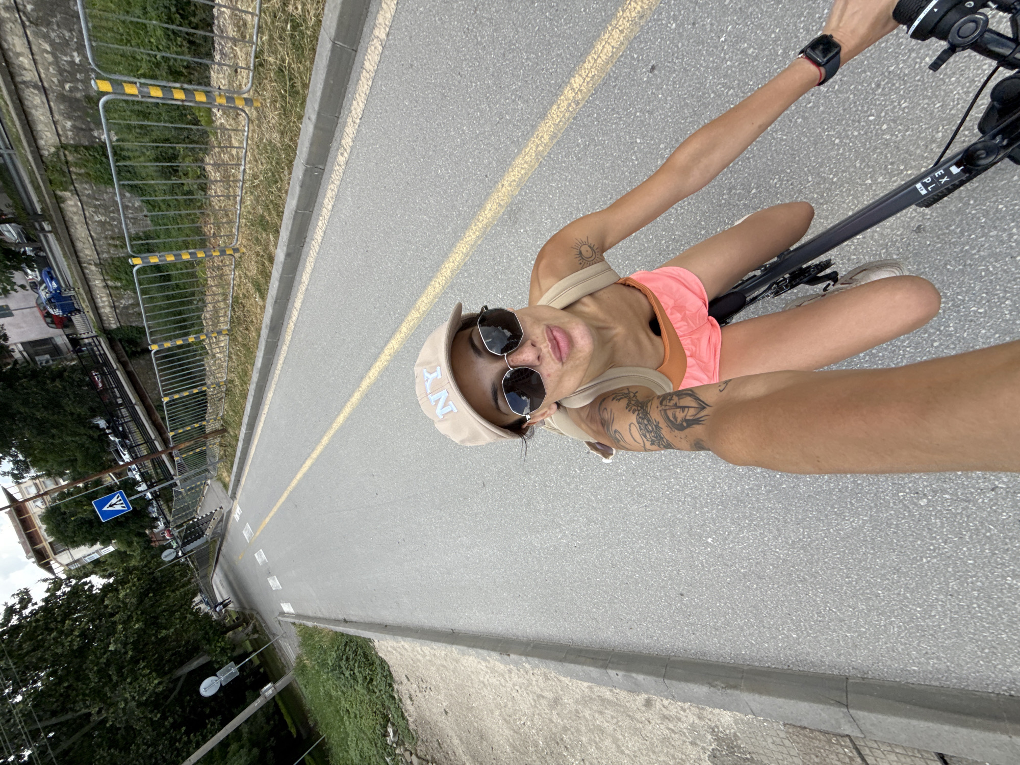 A woman with a tan complexion, wearing a tan baseball cap with "NY" embroidered on it, sunglasses, an orange tank top, and pink shorts, rides a bicycle on a paved road with a yellow lane divider. She has tattoos on her left arm and a sun tattoo on her right arm. Her right hand, wearing a smartwatch, is holding the handlebars. The road appears to be a dedicated bike path, bordered by a concrete curb and a metal fence. In the background, there are trees, a building, and parked cars. There is a pedestrian crossing marked with white stripes on the road. A blue pedestrian crossing sign is visible. The location is the city of Pleven, Bulgaria, near a small bridge over a stream or canal.