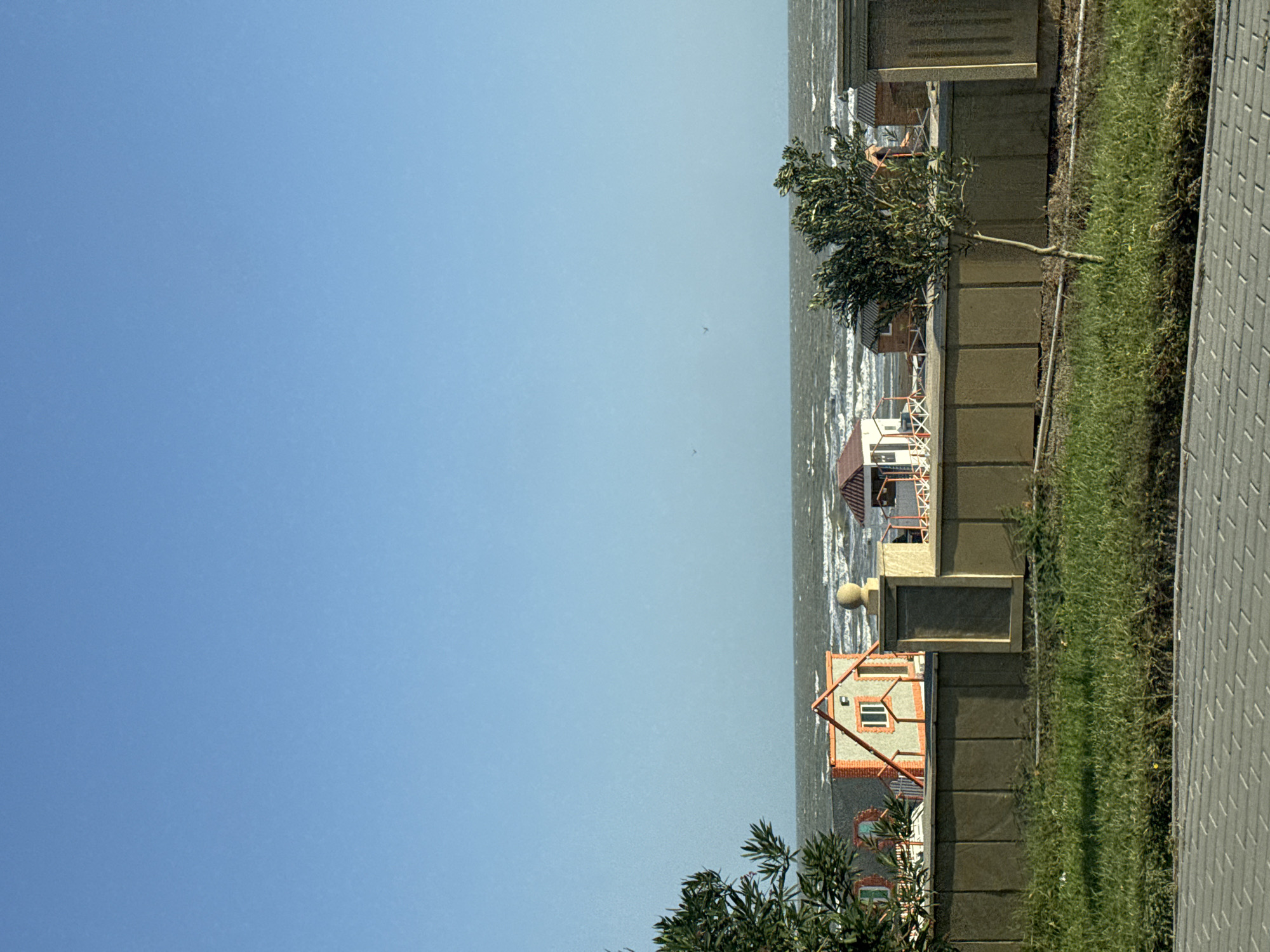 The image depicts a coastal scene with a clear blue sky. In the background, the sea is visible with moderate waves. Along the shore, there are several small, colorful structures that appear to be beach houses or changing rooms. In the foreground, a low, tan-colored wall with rectangular panels runs horizontally, separating the beach area from a strip of green grass. A small, slender tree stands to the right of the wall, and to the far left, a portion of a leafy tree is visible. The ground in front of the wall is covered with grass, and further in the foreground, a paved area with interlocking tiles is present. The overall scene suggests a tranquil day at a coastal resort or residential area.