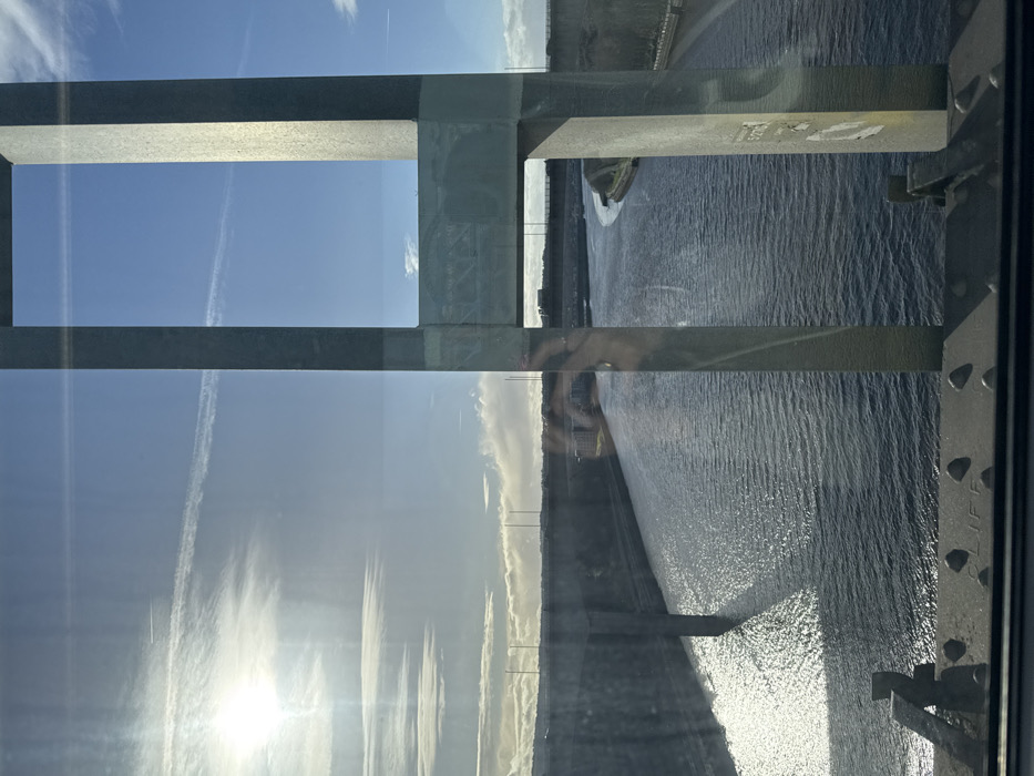 A partial view through a framed window reveals a sky with bright sunlight and wispy clouds on the left, contrasted by a body of water on the right. The water glistens in the sunlight, with a visible shoreline. Structural elements of a bridge or building frame the scene, with metal beams and supports visible. There are reflections in the glass, including hints of a person.