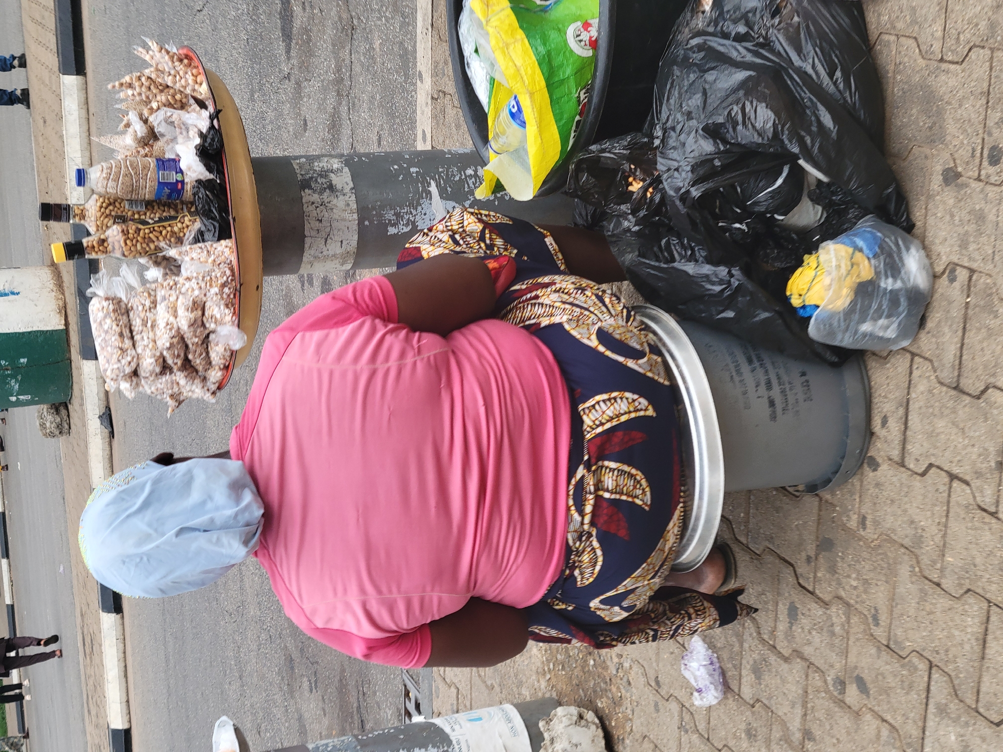 In Abuja, Nigeria, a street vendor sits on a gray bucket next to a light pole. The vendor is wearing a pink shirt, a patterned navy blue and brown skirt, and a light blue head covering. Next to the vendor is a black trash can filled with black plastic trash bags and a yellow plastic bag. In front of the vendor and attached to the pole is a wide dish filled with bags of peanuts and bottled nuts. The vendor is situated on a paved sidewalk near a street. Pedestrians are visible in the background on the sidewalk.