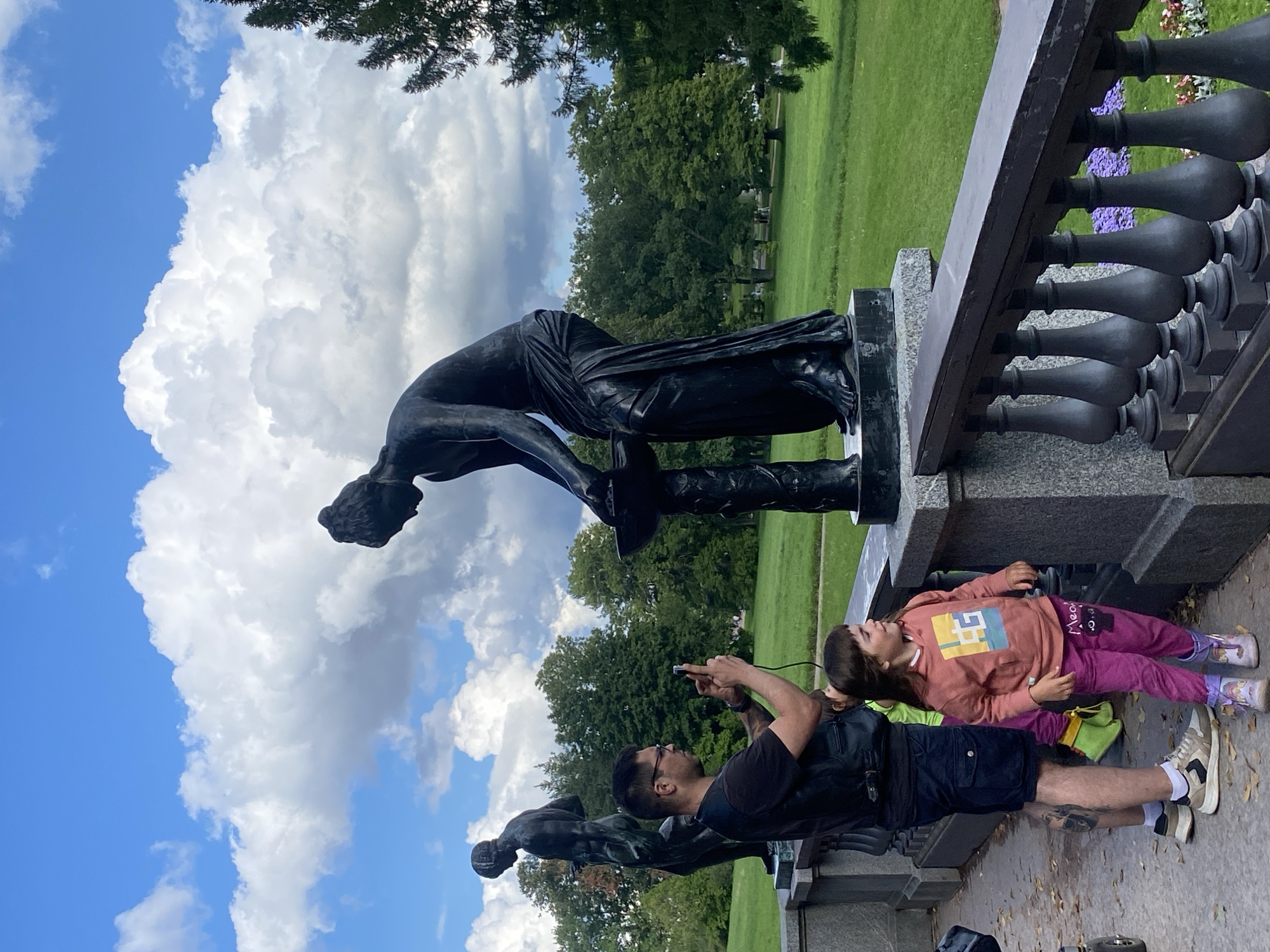 In the Catherine Park in Pushkin, Russia, two bronze statues of women are on display. One statue depicts a woman with her head bowed as she works with a basin on a pedestal. Another statue of a woman stands in the background. A man with a black vest over a black t-shirt and dark shorts is standing to the left of the first statue, holding up a smartphone as if to take a picture. He is wearing glasses and has tattoos on his legs. A young girl with a pink hooded sweatshirt and pink pants is standing next to the man. Her head is turned to the right, and she is looking upwards. A few autumn leaves are scattered on the stone walkway. The park features a green lawn and mature trees. The sky is blue with large, white, puffy clouds.