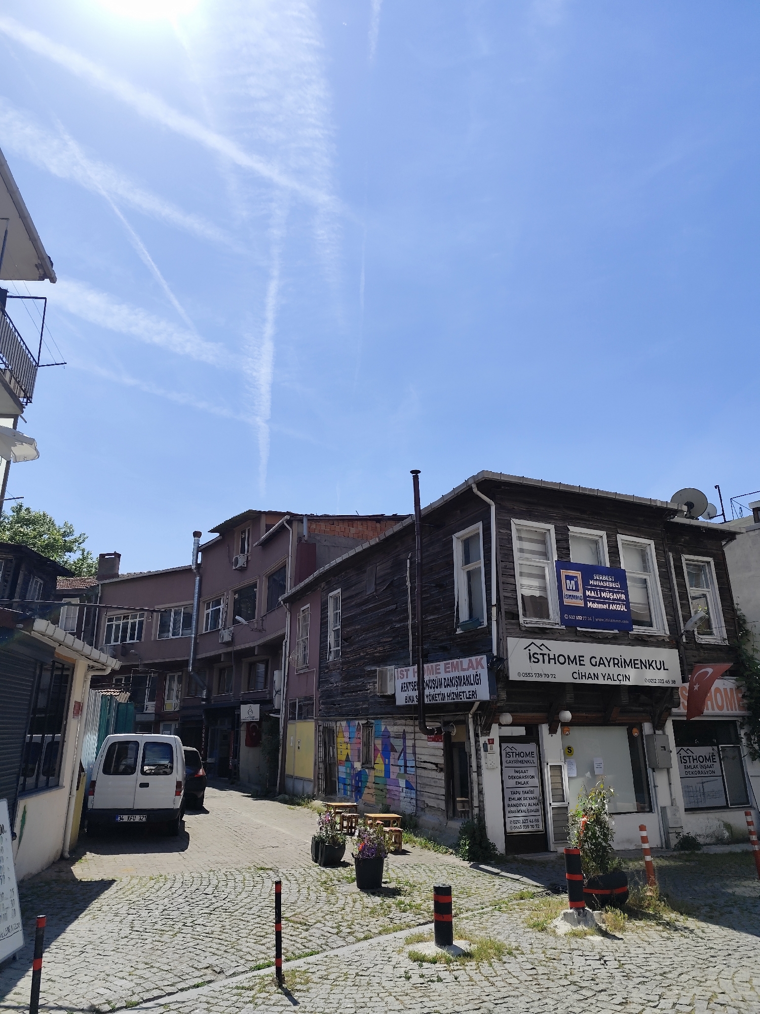 This image captures a bright, sunny street scene in what appears to be an older, urban neighborhood, likely in Sarıyer, Turkey, as indicated by the location context and Turkish text. The time of day is clearly midday or early afternoon, with strong sunlight casting shadows and a clear blue sky overhead, crisscrossed by several prominent contrails forming an "X" shape.
The scene features a narrow, cobbled street with buildings on both sides. On the right, a weathered, two-story wooden building with multiple windows dominates the foreground. This building houses several businesses, prominently displaying multiple signs for "İSTHOME GAYRİMENKUL" (ISTHOME REAL ESTATE) offering services like urban transformation consultancy, property management, deed follow-up, and inheritance transfer transactions. A sign for "SERBEST MUHASEBECİ MALİ MÜŞAVİR Mehmet AKGÜL" (Independent Accountant Financial Advisor Mehmet AKGÜL) is also visible on the upper floor, along with phone numbers and a website. A Turkish flag hangs outside one of the shopfronts.
Further down the street, other buildings with varying facades, some painted brown or purplish-grey, line the path. A white van with the license plate "34 KFU 321" (indicating Istanbul) is parked on the left side of the street, alongside a dark car. No people are visible, suggesting a quiet moment in the neighborhood. Elements like satellite dishes on rooftops, air conditioning units, and bollards on the cobblestones add to the urban texture. Outside the main wooden building, there are flower pots, tables, and chairs, hinting at a small outdoor seating area. On the far left, a small sign reads "LEMON 200".