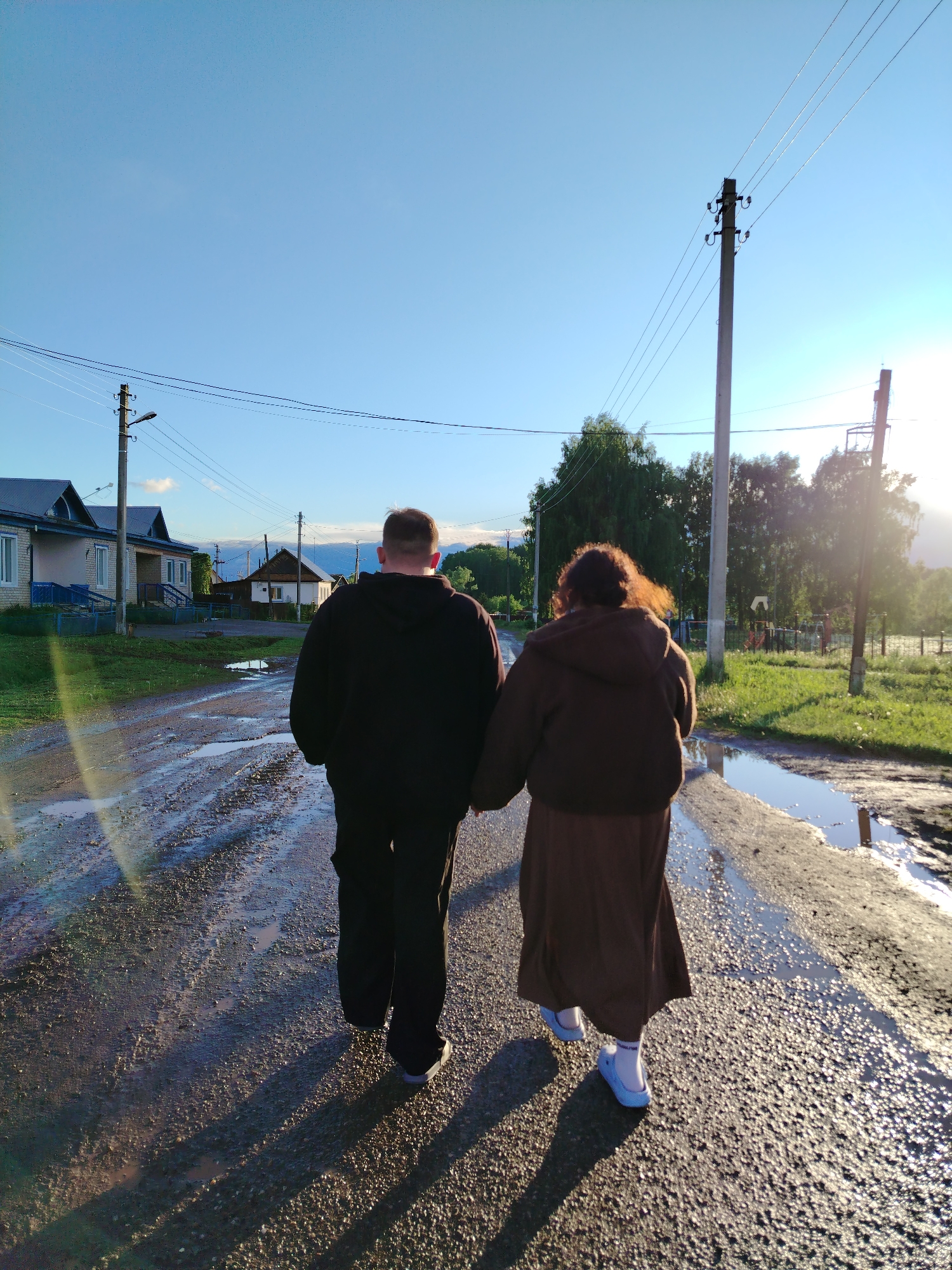 Two people are walking hand-in-hand on a wet road. The person on the left is wearing a dark hooded sweatshirt and dark pants. The person on the right is wearing a brown hooded jacket and a long brown skirt, paired with light blue shoes. The road they are walking on is wet with puddles, reflecting the sky. Buildings and power lines are visible in the background, suggesting a rural or suburban environment. Greenery is present on either side of the road. The image was taken in Гожан, Russia.