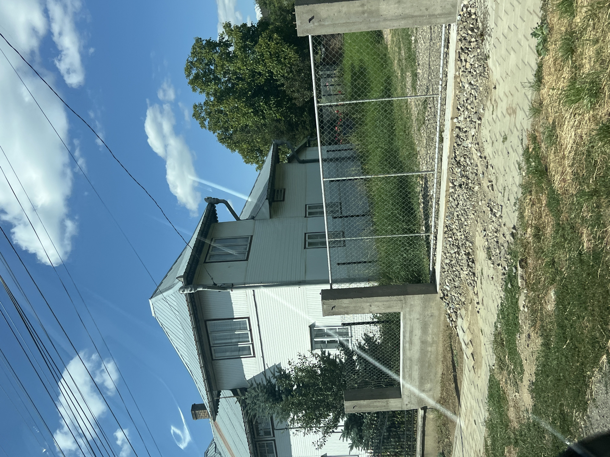 The image captures a white, two-story house with a metal roof, situated in Capu Codrului, Romania. A chain-link fence with concrete posts encloses the property, and a paved walkway with decorative elements leads to the entrance. A small evergreen tree stands in front of the house, and a larger deciduous tree is visible in the background. The sky is blue with scattered white clouds, and power lines are strung across the top of the frame. The overall scene depicts a residential area on a sunny day.