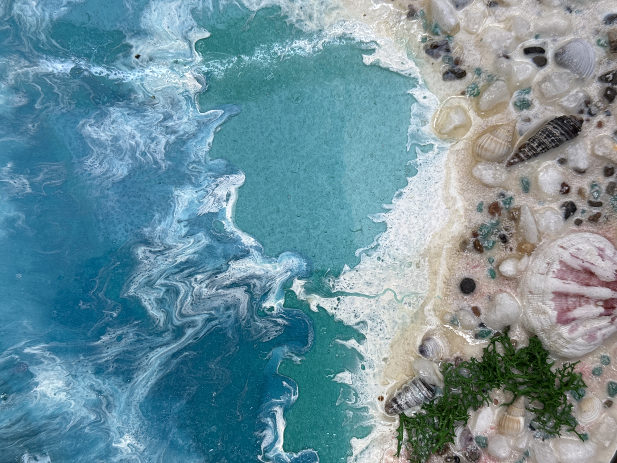 A close-up, overhead shot captures a detailed resin art piece depicting a beach and ocean scene. The upper portion of the artwork features swirling patterns of blues and white, mimicking ocean waves and foam. Transitioning downwards, a turquoise area represents shallow water, bordered by a frothy white "wave" made of resin. The bottom of the image showcases a sandy beach adorned with various shells, small stones, and a cluster of green faux seaweed. The overall impression is one of a vibrant, miniature coastal landscape.