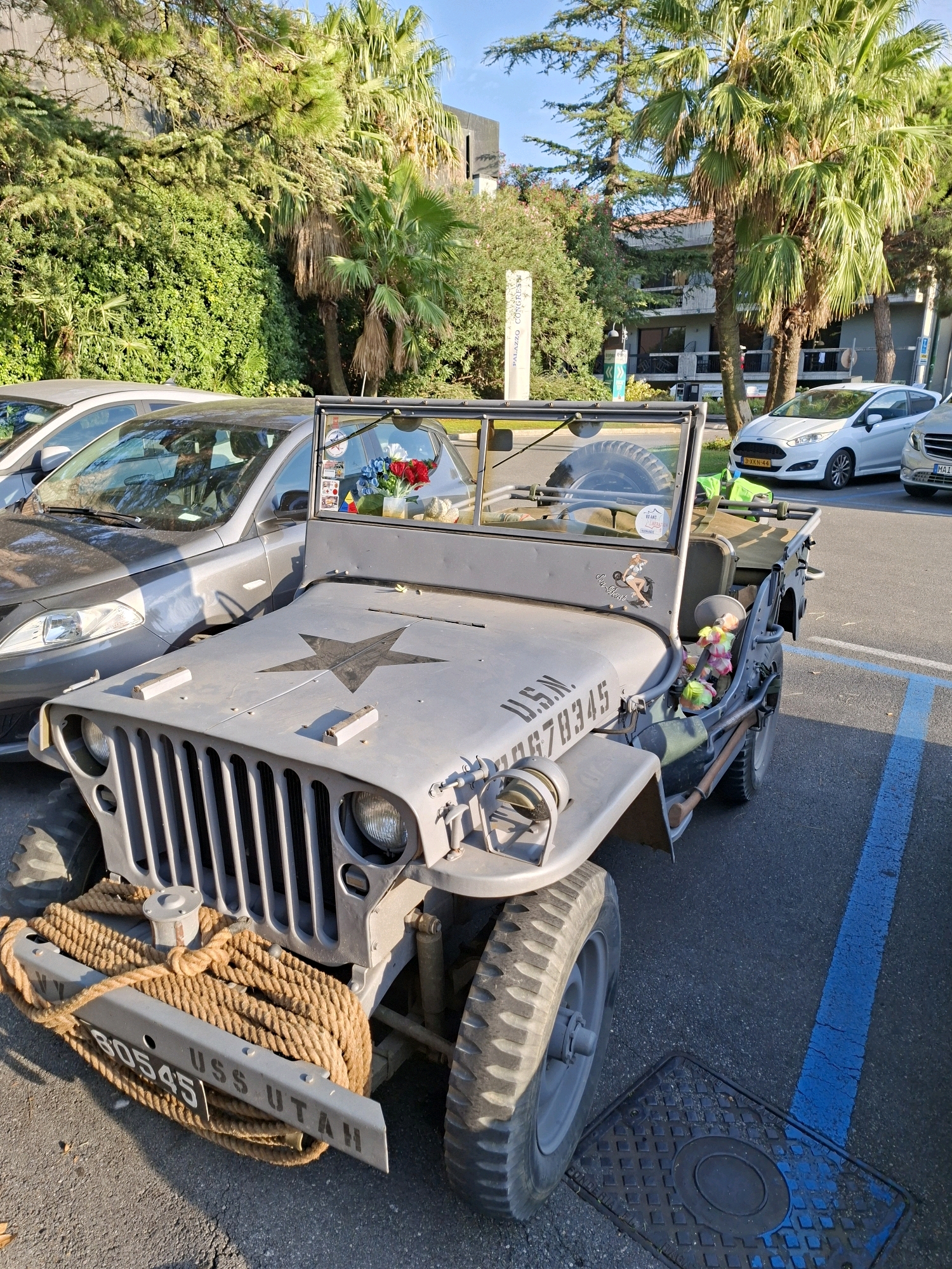 The image shows a vintage grey Jeep parked outdoors in Sirmione, Italy. The Jeep has a large black star on its hood and the designation "U.S.N. 10678345" painted on its side. A thick rope is wrapped around the front bumper, which also has "USS UTAH" and "B0545" written on it. The Jeep's windshield is down, and a small bouquet of artificial flowers, including red and blue roses, is visible inside. There is a pin-up sticker of a woman on the driver's side of the Jeep.
The Jeep is parked next to a dark grey sedan and a white Ford Fiesta. In the background, palm trees and other lush green foliage are visible, suggesting a warm climate. A building with windows and balconies can be seen behind the trees. A blue painted line on the asphalt indicates a parking space. The scene appears to be a parking area, possibly near a public building or tourist attraction, given the presence of the Palazzo Congressi sign partially visible in the distance. The lighting indicates it is daytime, with sunlight casting shadows. There are no people directly visible in the image, but the presence of cars and the setting suggests a populated area.