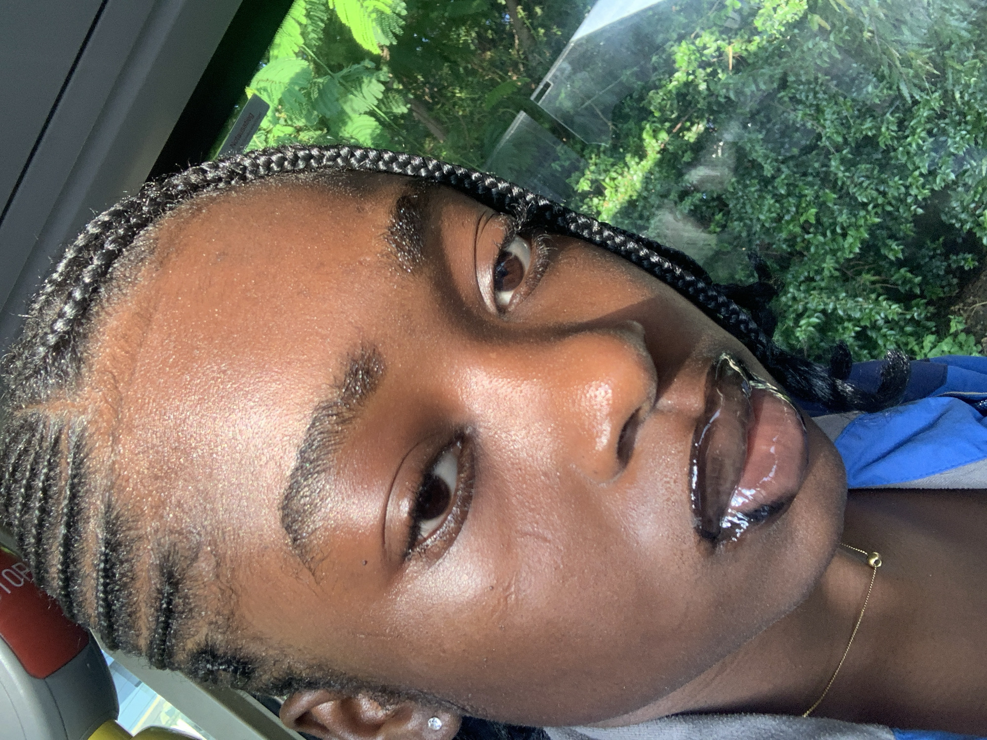 A young Black person with braided hair and clear, dewy skin poses for a close-up selfie. They are wearing a blue jacket and a thin gold necklace. The person's lips are glossy and reflecting light. The background shows a blur of green foliage, suggesting they are outdoors or near a window with a view of trees. The selfie is taken from a low angle, tilted to the side.