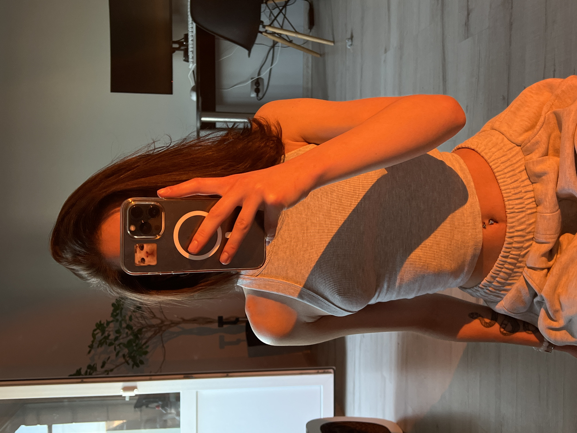 The image depicts a young woman taking a selfie in what appears to be an indoor setting, possibly a living room or studio apartment. She is seated on the floor, holding a smartphone in her right hand to capture the reflection. The woman has long, dark hair and is wearing a light grey ribbed tank top and light grey sweatpants. A navel piercing is visible. Her left arm has a tattoo of a snake. The lighting in the room is warm and orange-toned, suggesting it might be sunset or artificial lighting. In the background, a potted plant, a desk with a computer monitor and keyboard, and a chair are visible. The floor is wooden. The image does not contain any specific identifying features or context that would allow for its location to be determined in Ternopil, Ukraine.
