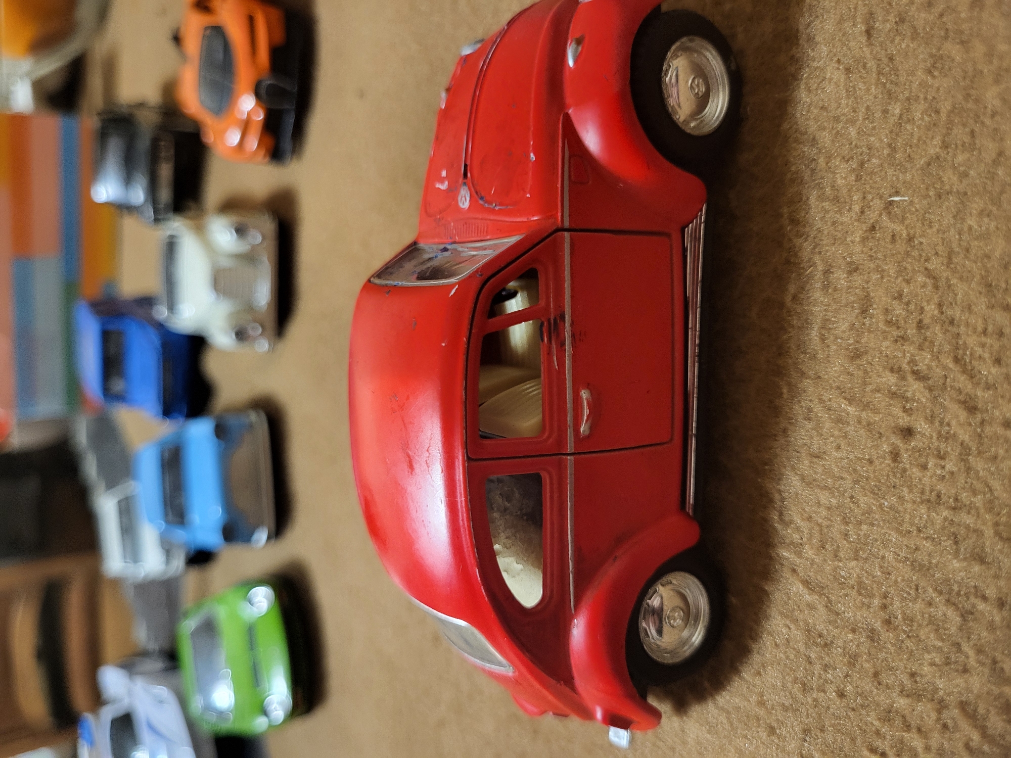 The image shows a collection of toy cars on a brown, textured surface. A red toy car, resembling a Citroen 2CV, dominates the foreground. Behind it, a series of smaller toy cars are arranged in a line, exhibiting a variety of colors including orange, black, white, blue, and green. The image is likely indoors. The location in Yerevan, Armenia is not discernible from the visual information.